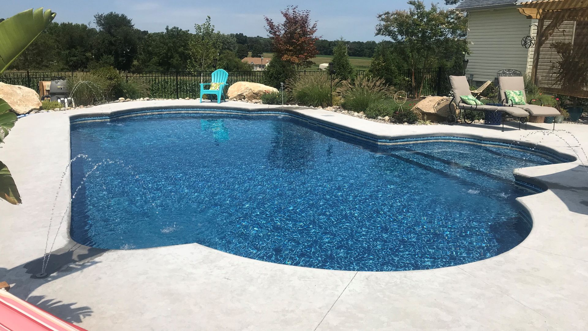 Pool with blue water and concrete patio, surrounded by rocks and landscaping, on a sunny day.