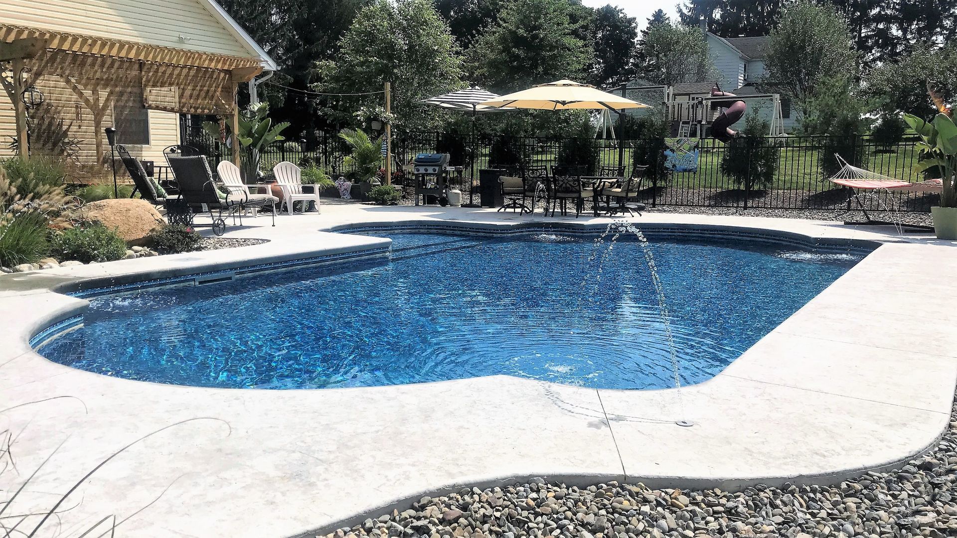Swimming pool with blue water, surrounded by concrete and landscaping. Deck and patio furniture visible.