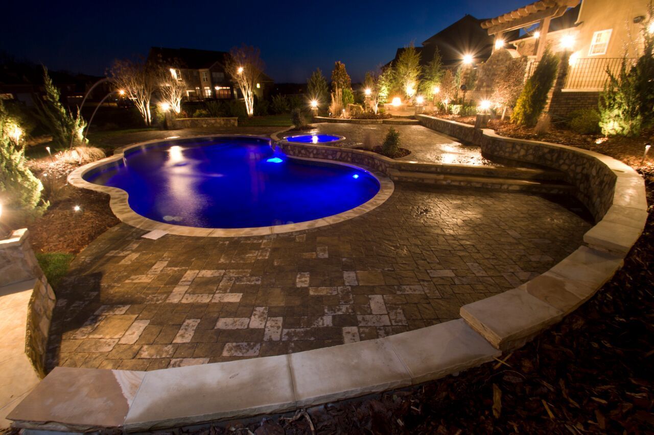 Night view of a backyard pool and patio, illuminated by warm lights. Blue pool water reflects the glow.