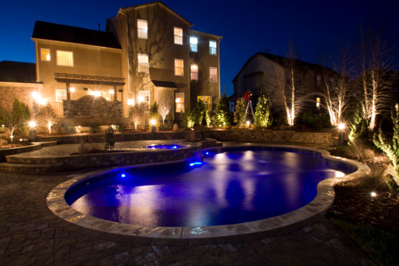Night view of a luxury home with a blue-lit pool, patio, and illuminated landscaping.