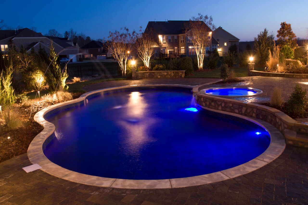Night view of a kidney-shaped pool with blue lights.  A hot tub and house are in the background, lit with outdoor lights.