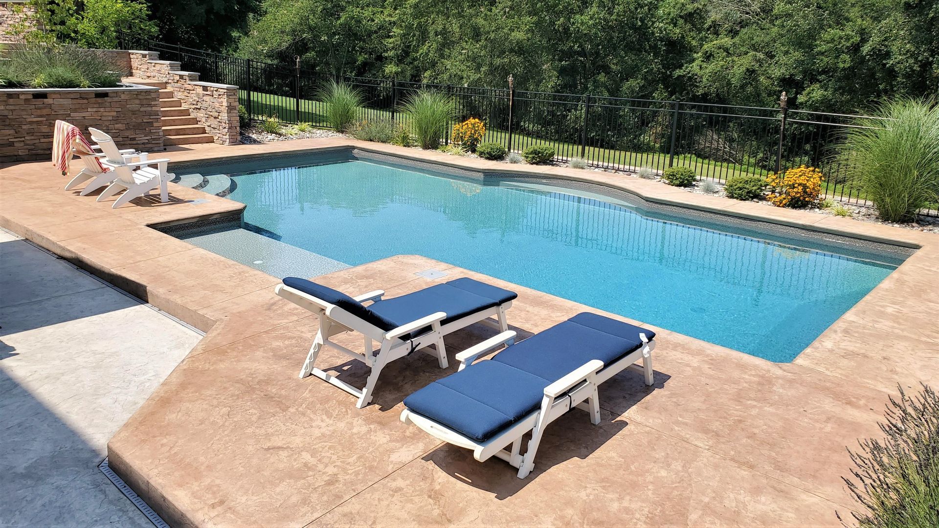 Swimming pool with two lounge chairs on a concrete patio. Steps lead into the blue water. Green foliage surrounds.