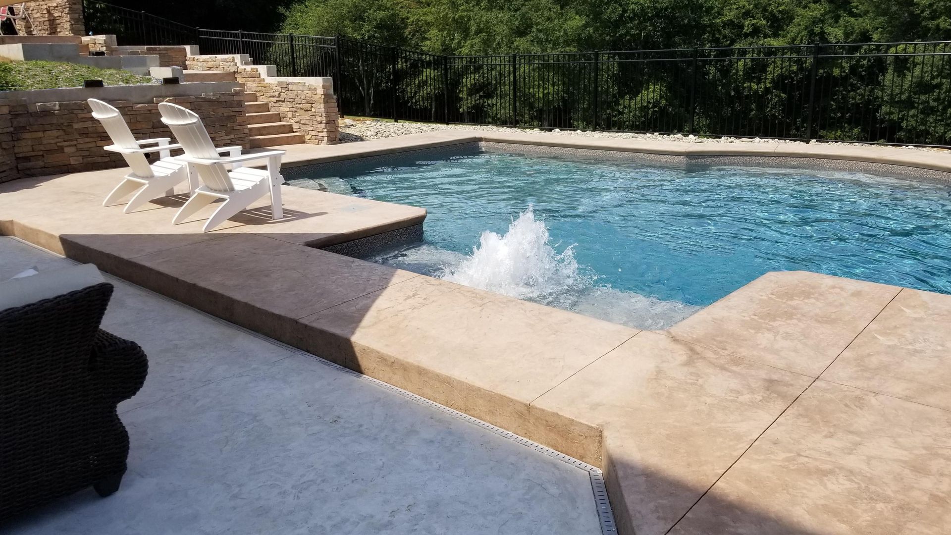 Pool with cascading water feature, lounge chairs, and stone patio. Blue water against brown and beige tones.
