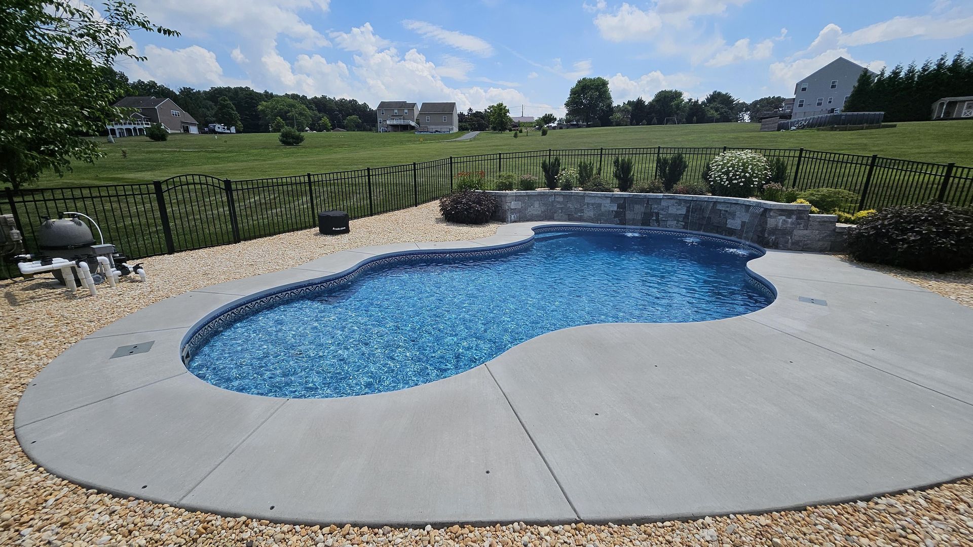 Swimming pool with a blue interior, surrounded by concrete and gravel, with a green lawn and houses in the background.