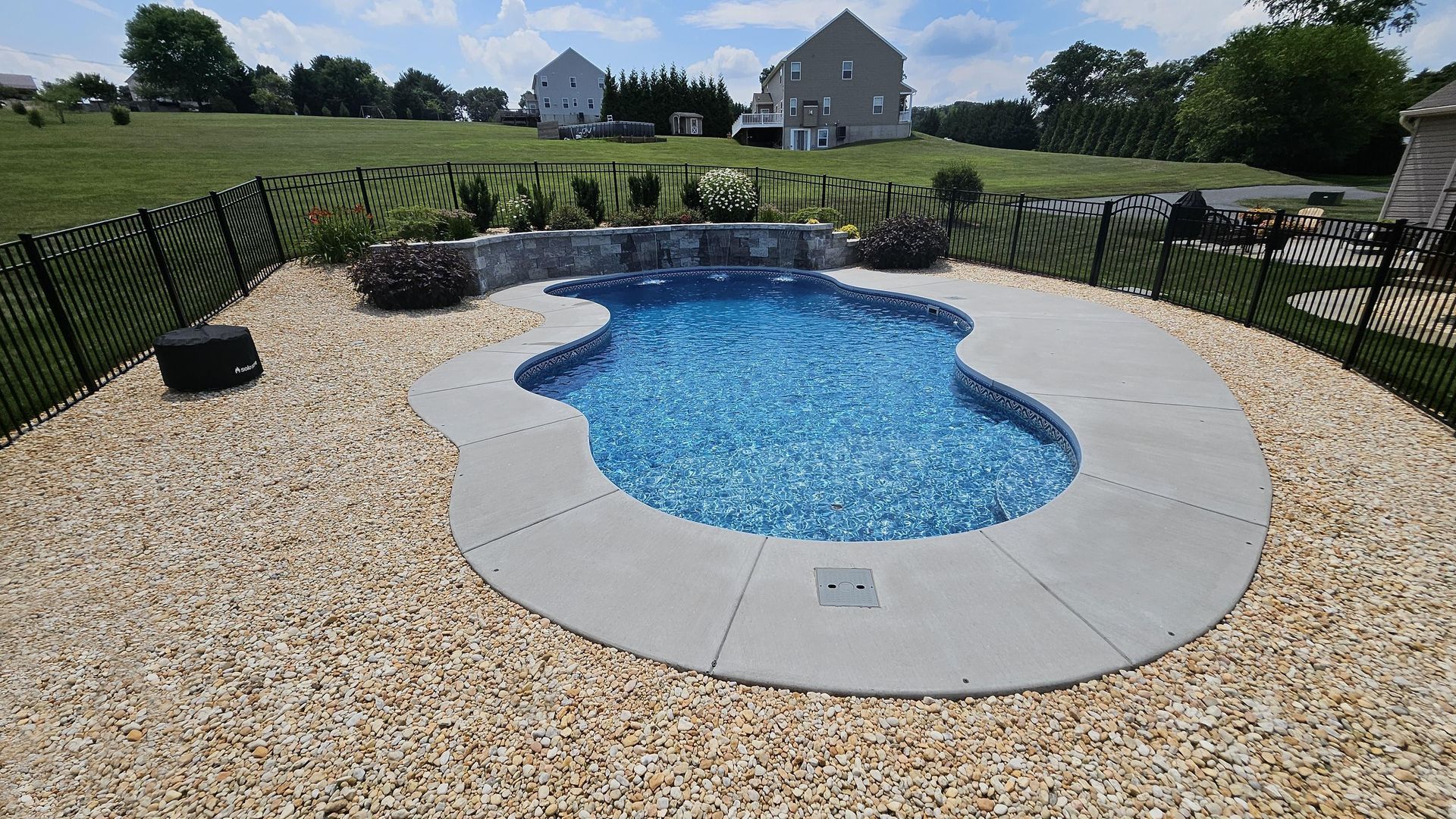 Small, freeform pool with blue tile surrounded by tan gravel and concrete, black fence.