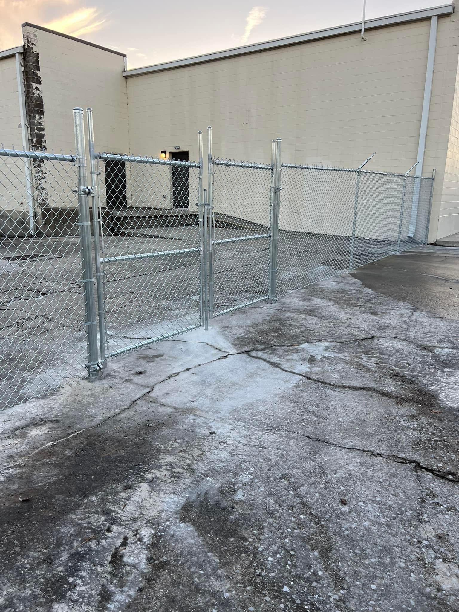 A chain-link fence with a closed gate stands in front of a beige building on a cracked, gravel-covered lot.