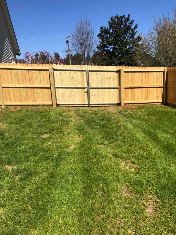 A wooden privacy fence with a central gate stands in a grassy backyard under a clear blue sky.