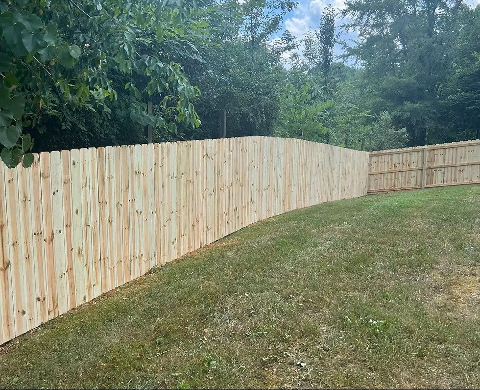 A new, light-colored wooden privacy fence stands on a grassy lawn with a tree line in the background.