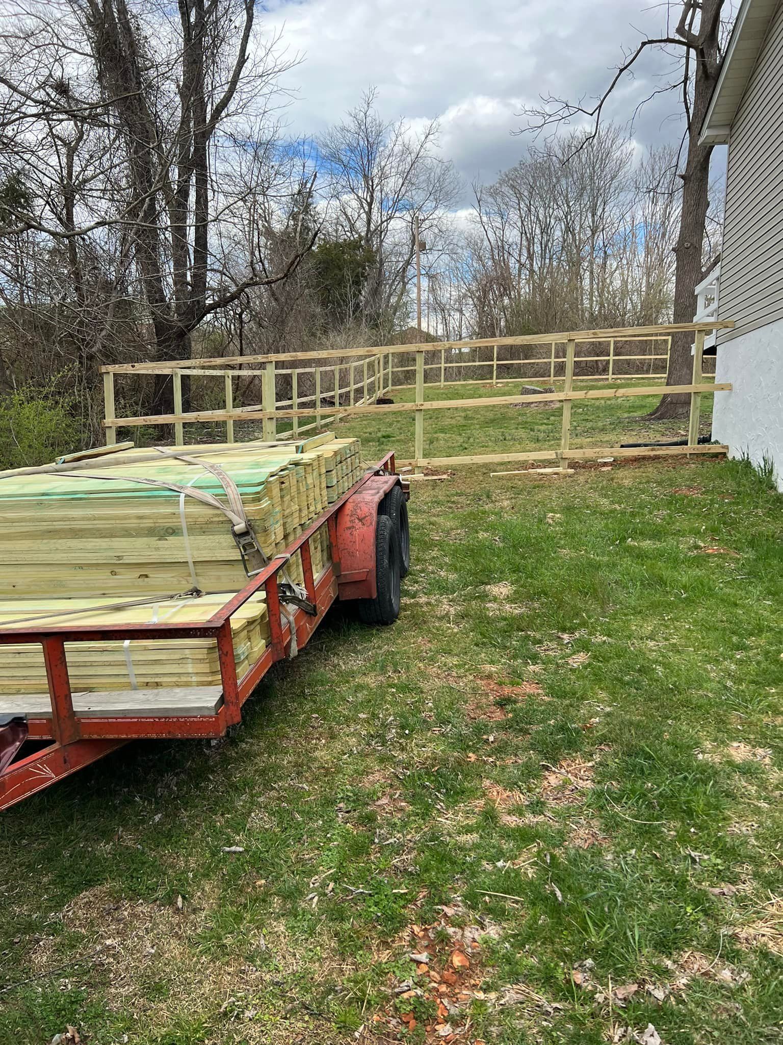 A red trailer loaded with lumber parked on a grassy yard next to a partially built wooden fence.