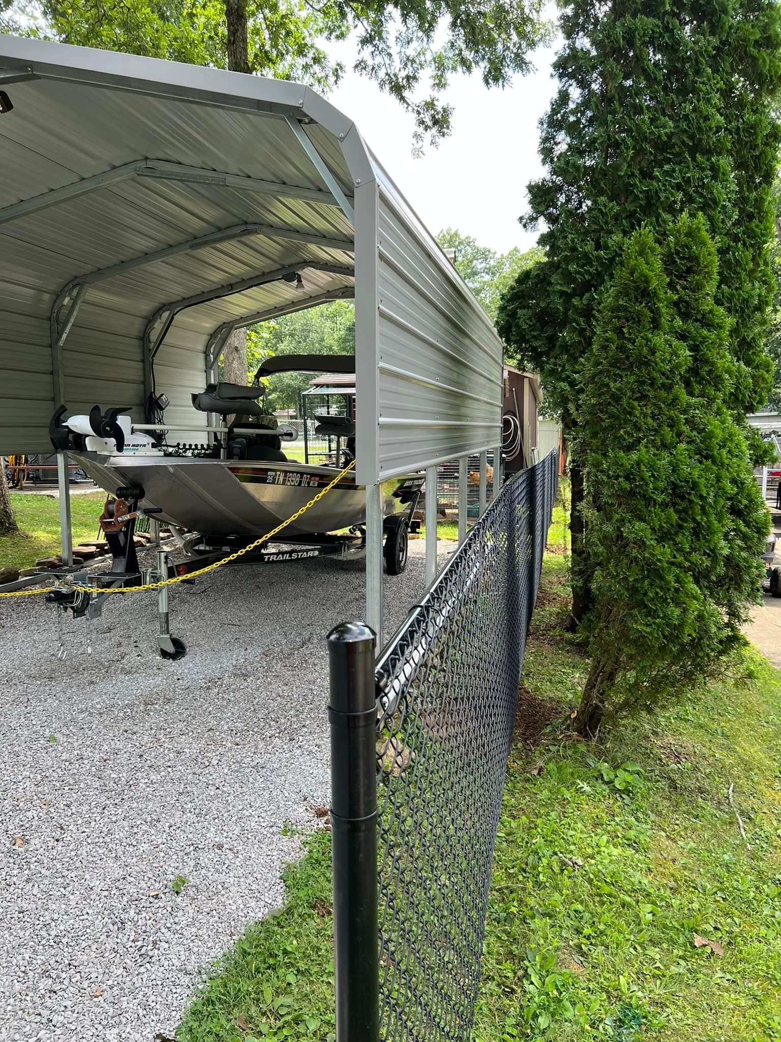 A metal carport sheltering a boat parked on a gravel surface behind a black chain-link fence, next to tall green trees.