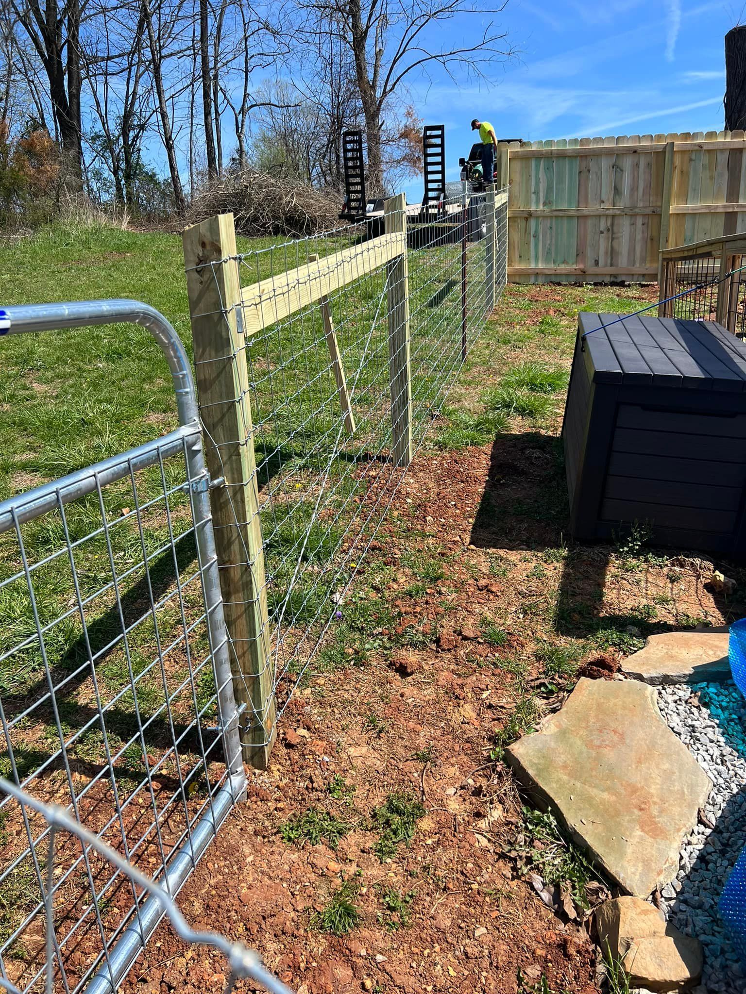 A wooden and wire fence separates a grassy yard from a mulch bed, next to a metal gate and a black deck box.