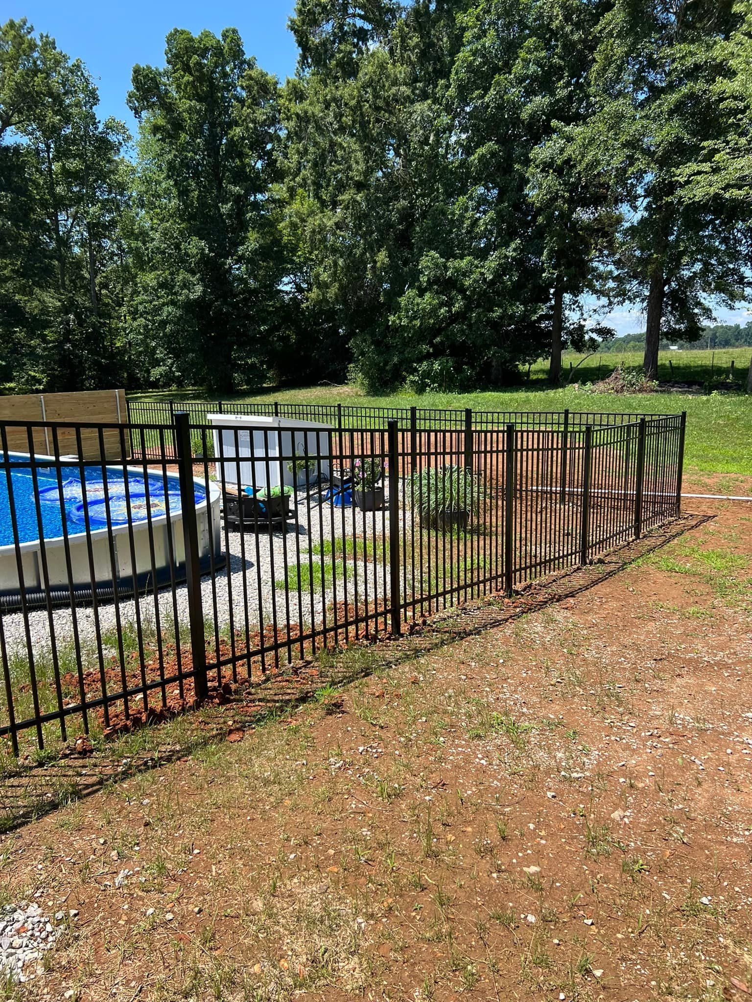A circular above-ground pool enclosed by a black metal fence on a grassy lot with trees in the background.