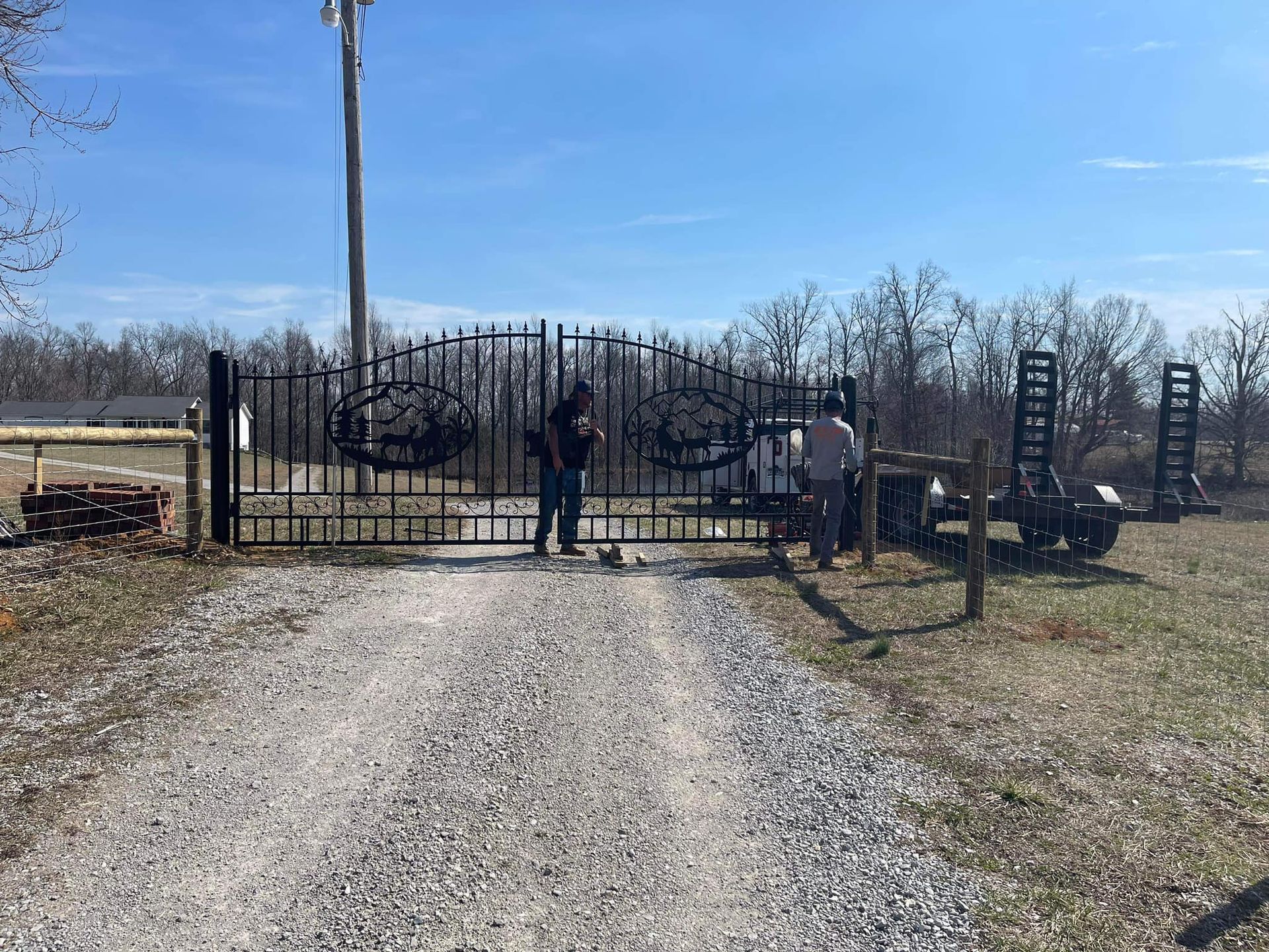 Two people stand near a decorative black metal gate at the entrance to a rural gravel driveway on a sunny day.
