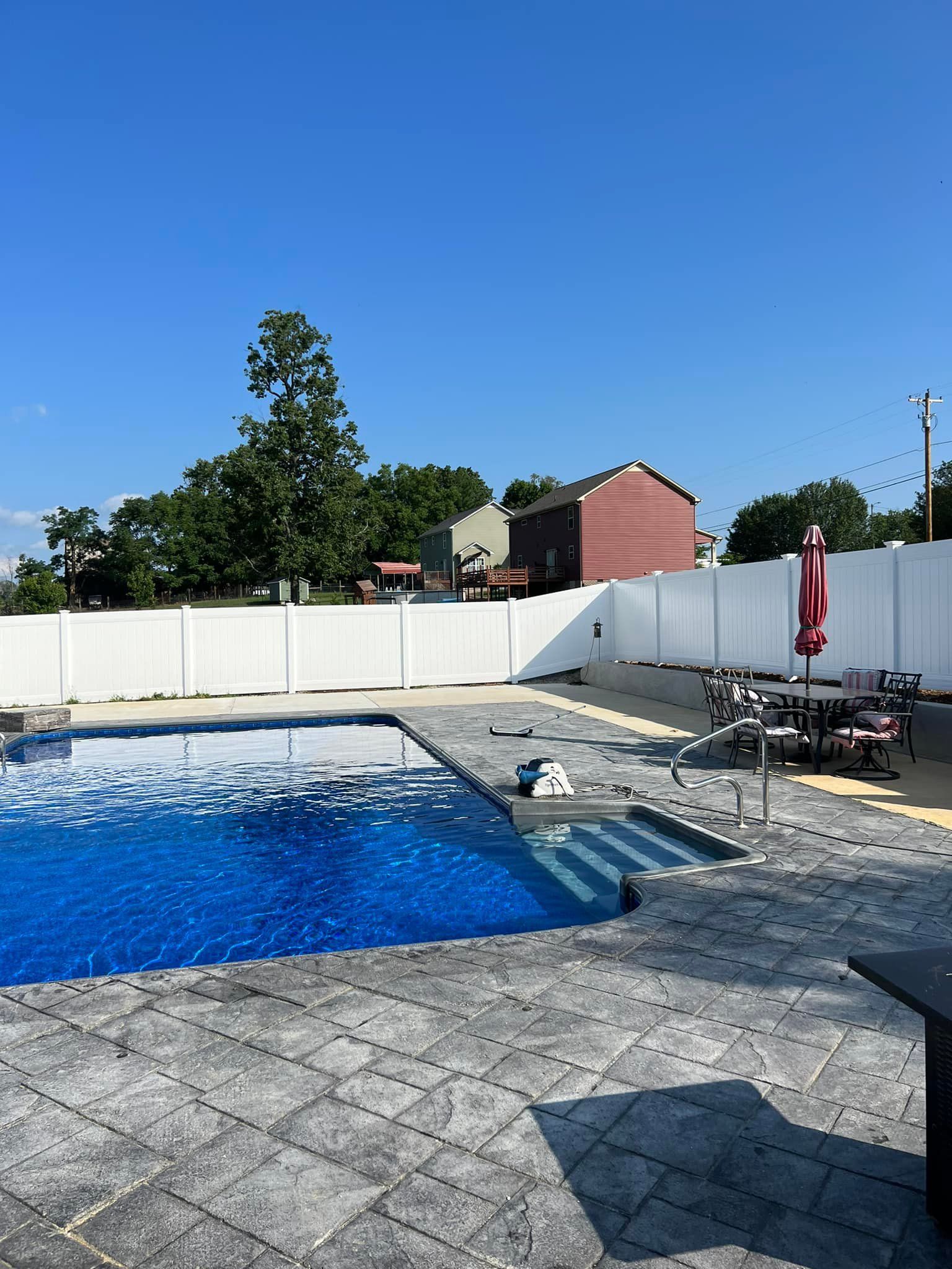 A backyard swimming pool with deep blue water, surrounded by gray stone pavers and a white vinyl fence on a sunny day.