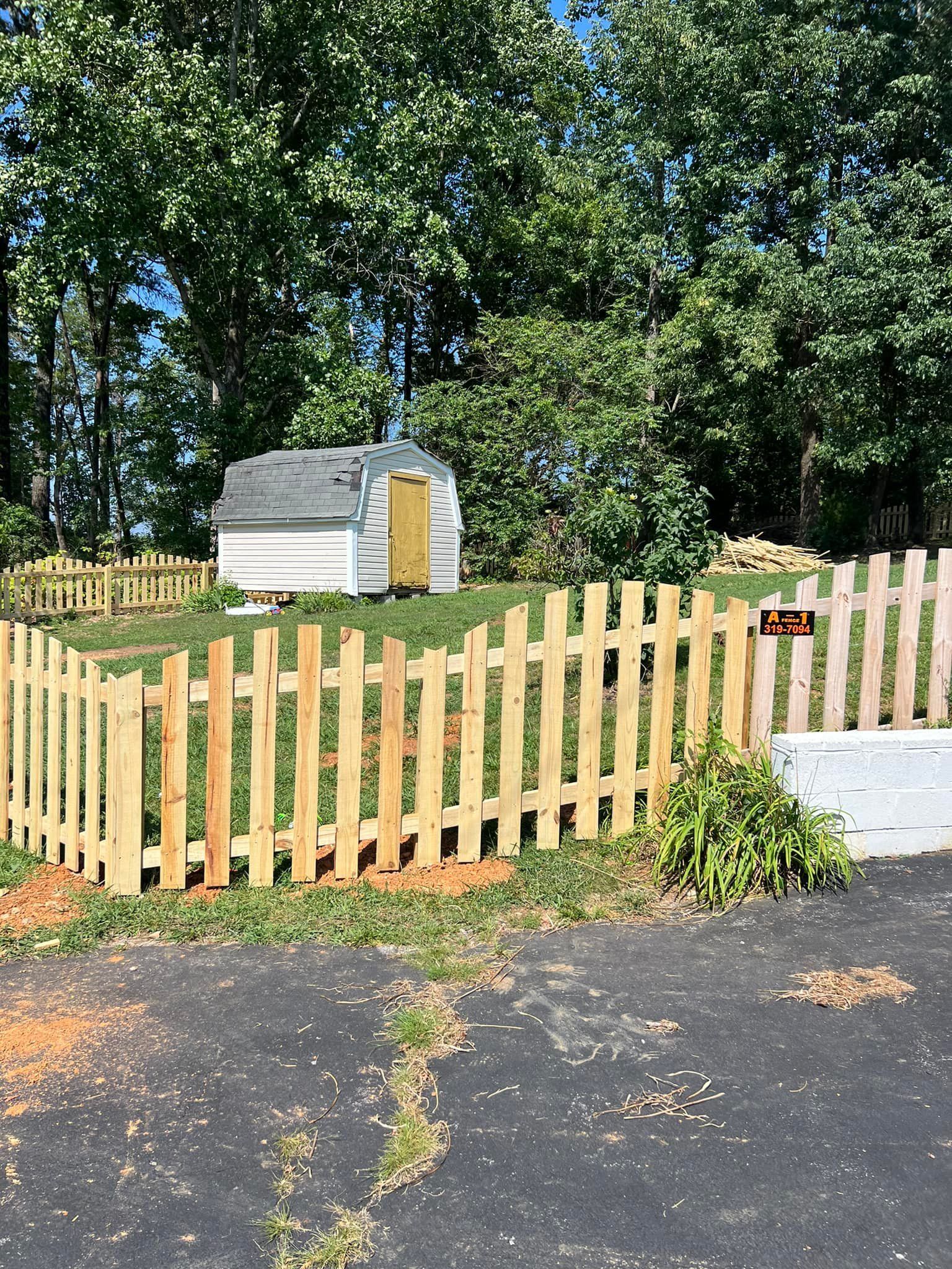 A white shed sits in a grassy yard behind a newly installed wooden picket fence, set against a backdrop of green trees.