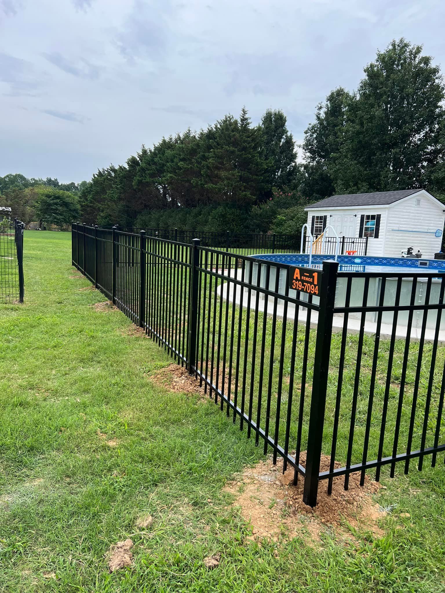 A black metal fence encloses an above-ground pool in a grassy backyard near a white shed under a cloudy sky.