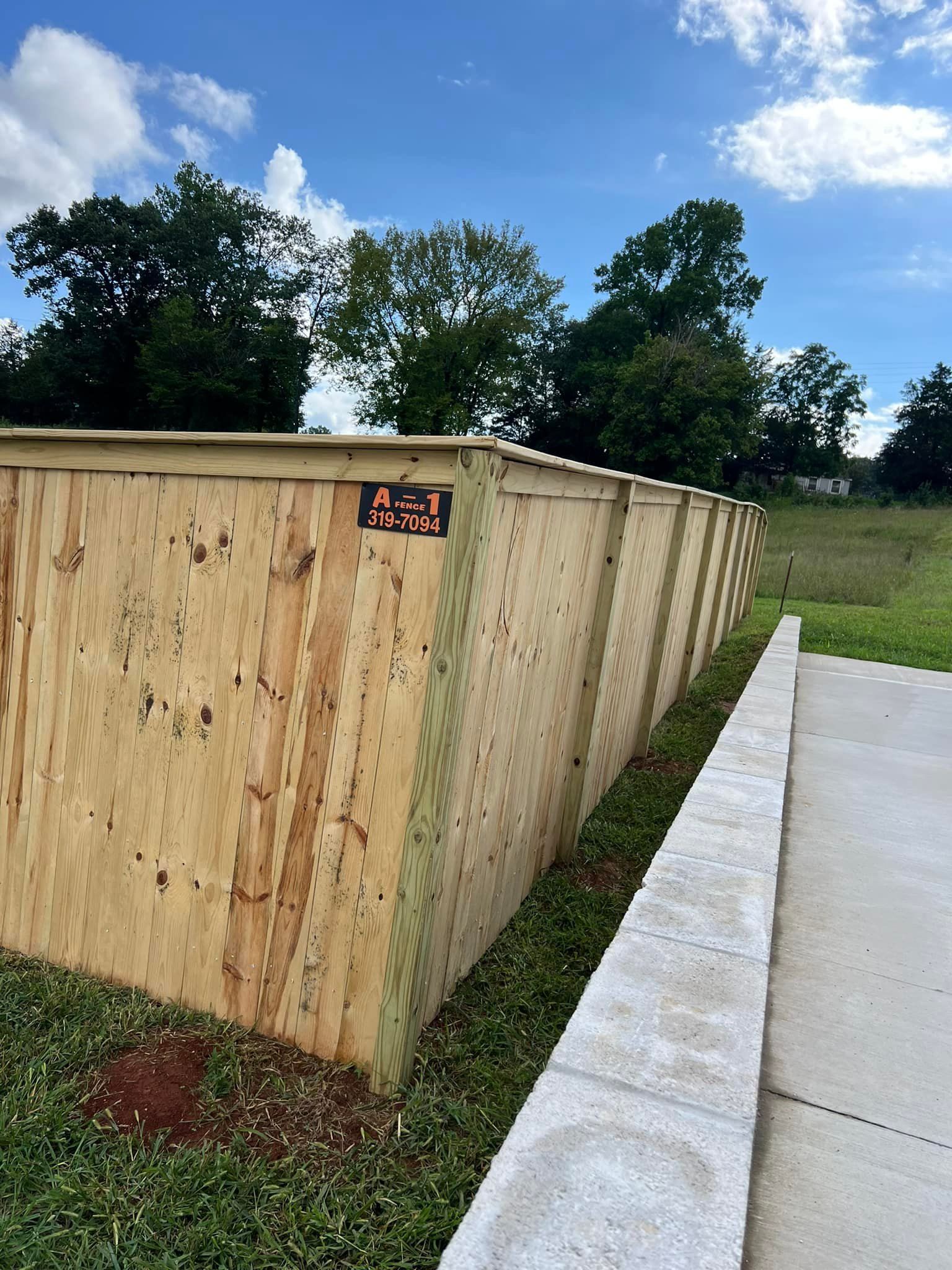 A long plywood construction wall stands next to a concrete sidewalk and a grass strip under a blue sky with clouds.