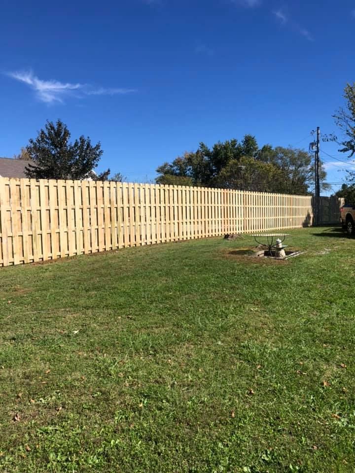 A new, light-colored wooden fence spans across a grassy backyard under a bright blue sky.
