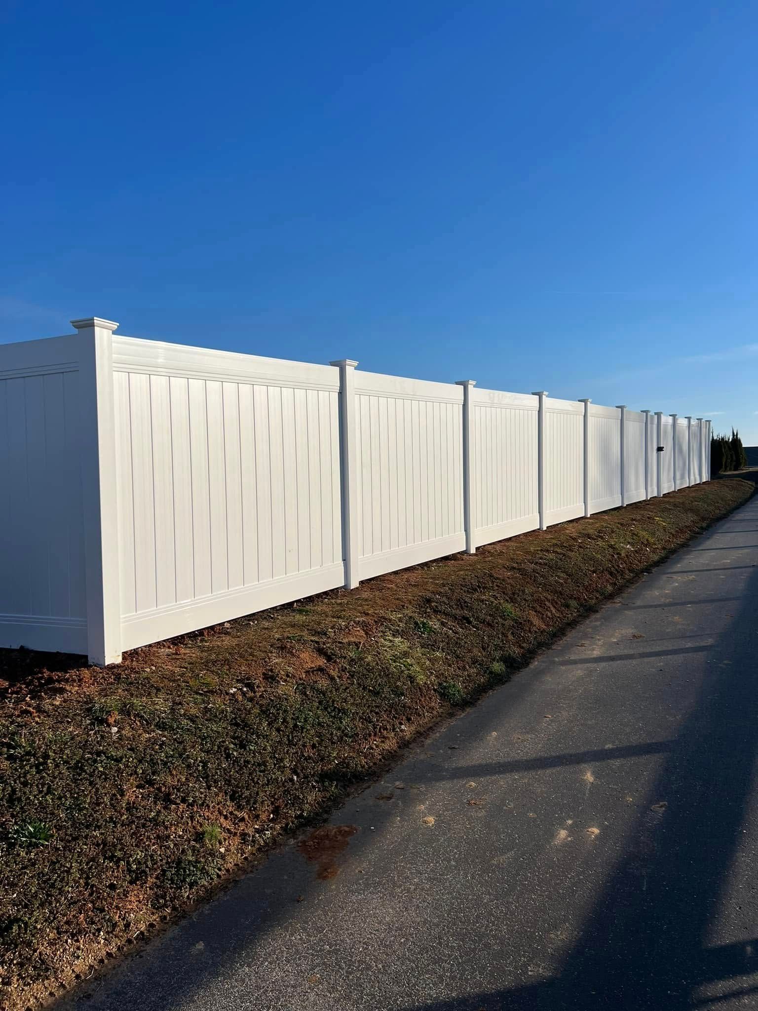A long white vinyl privacy fence stands next to a paved path and a patch of dry grass under a clear blue sky.