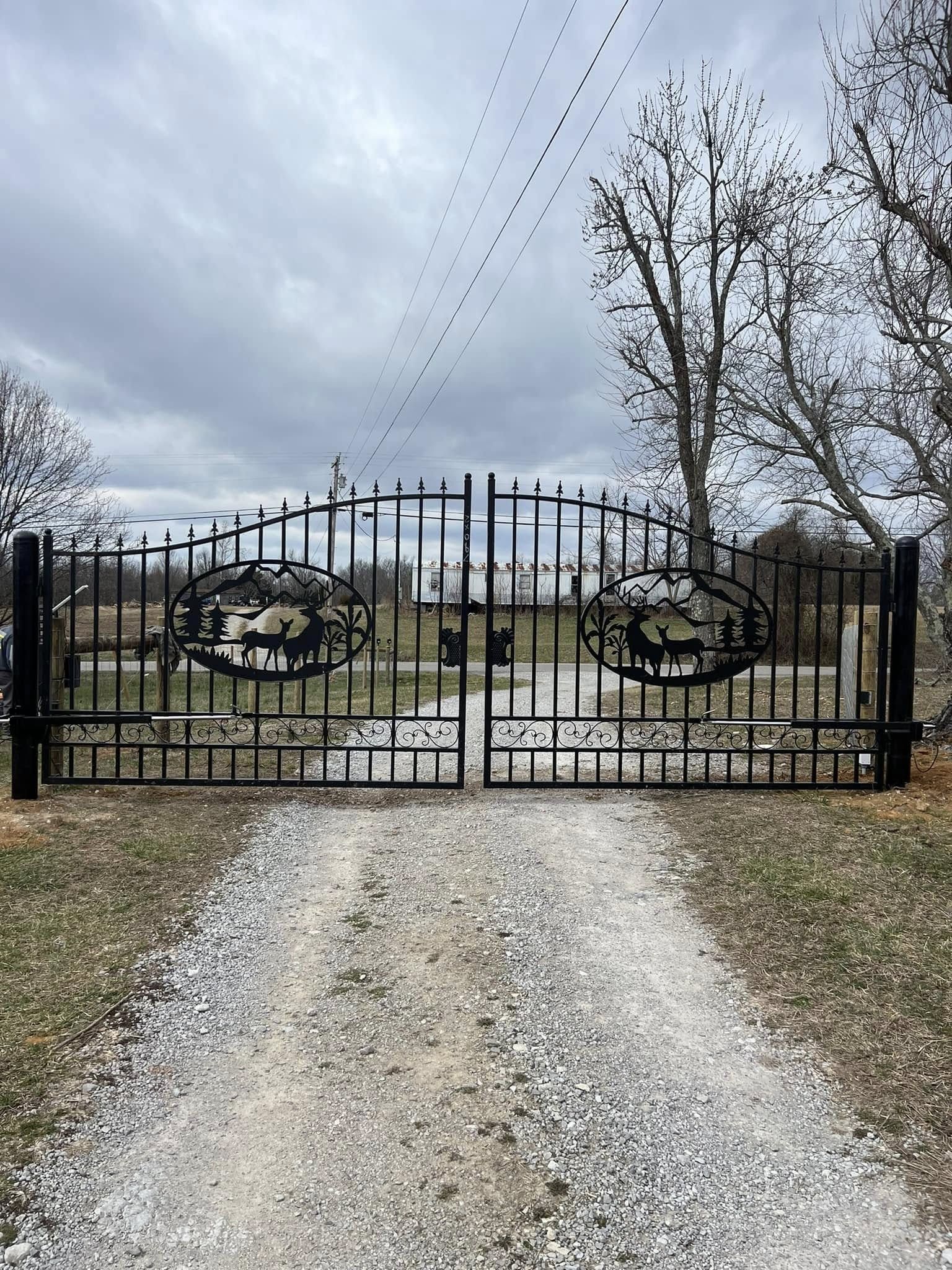 A black decorative metal gate with deer silhouettes in the center, spanning a gravel driveway under a cloudy sky.