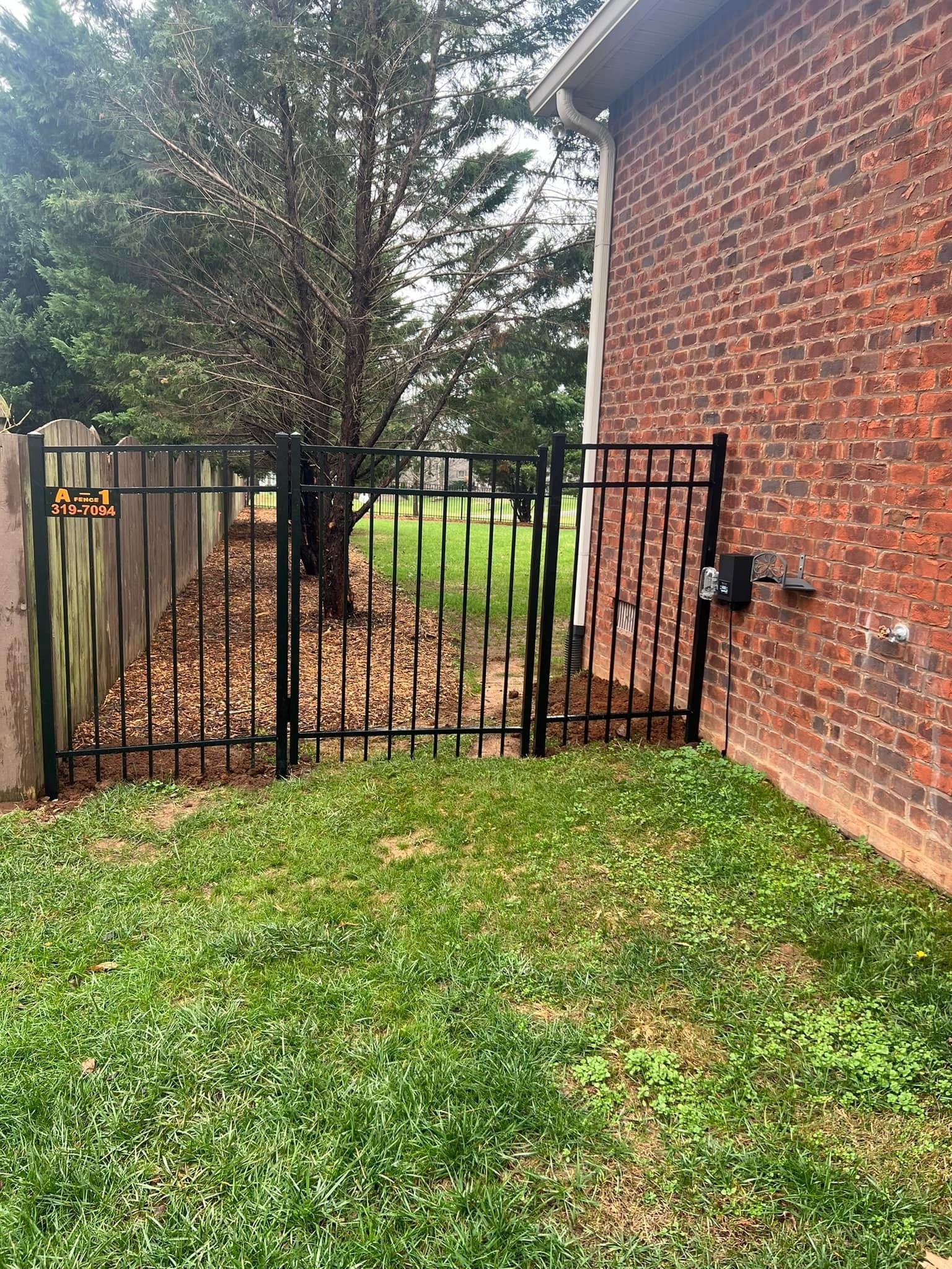 A black metal gate set between a brick house wall and a wooden fence, leading to a grassy yard with trees.