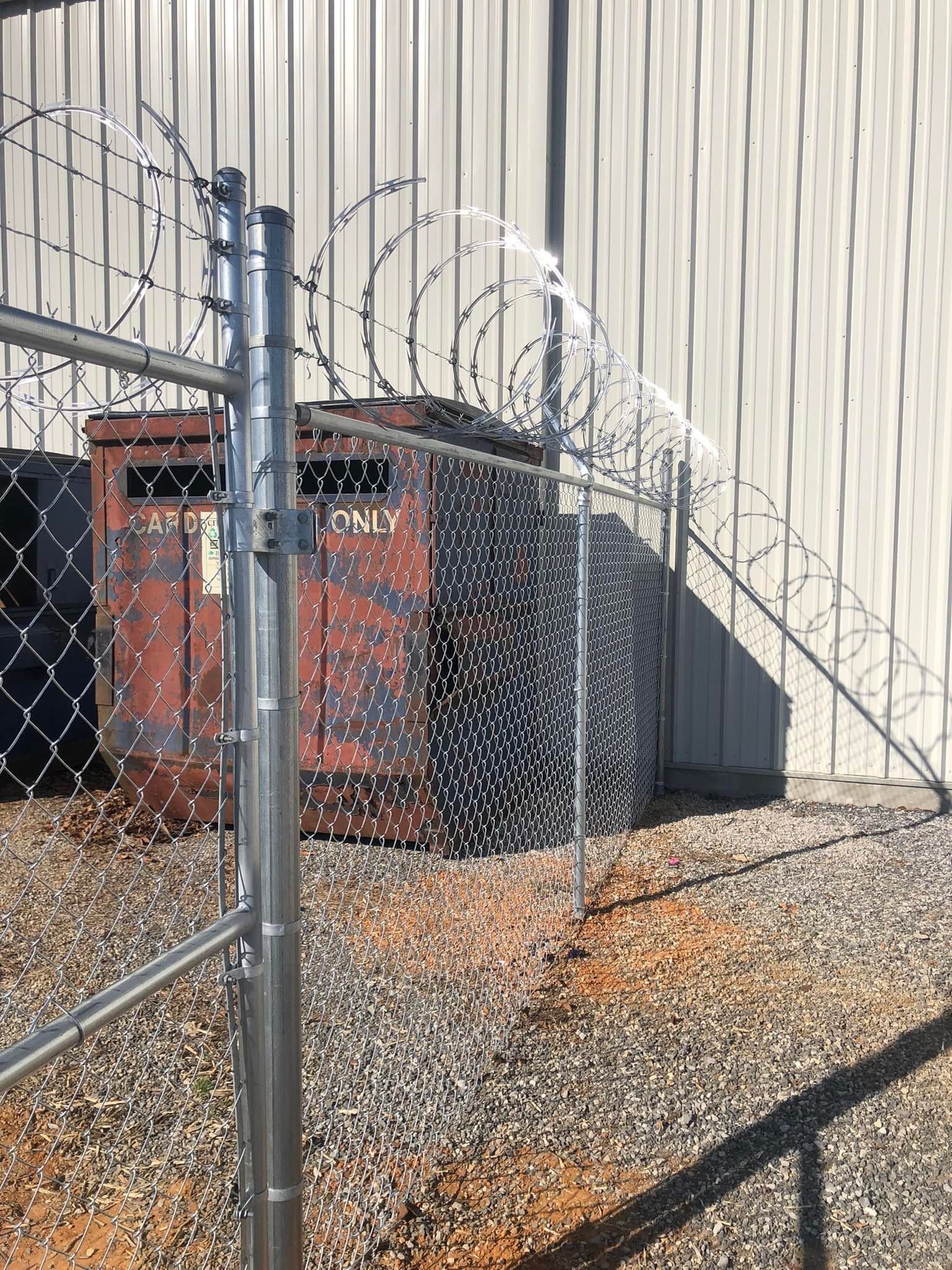 A chain-link fence topped with barbed wire stands in front of a reddish dumpster and black industrial equipment.