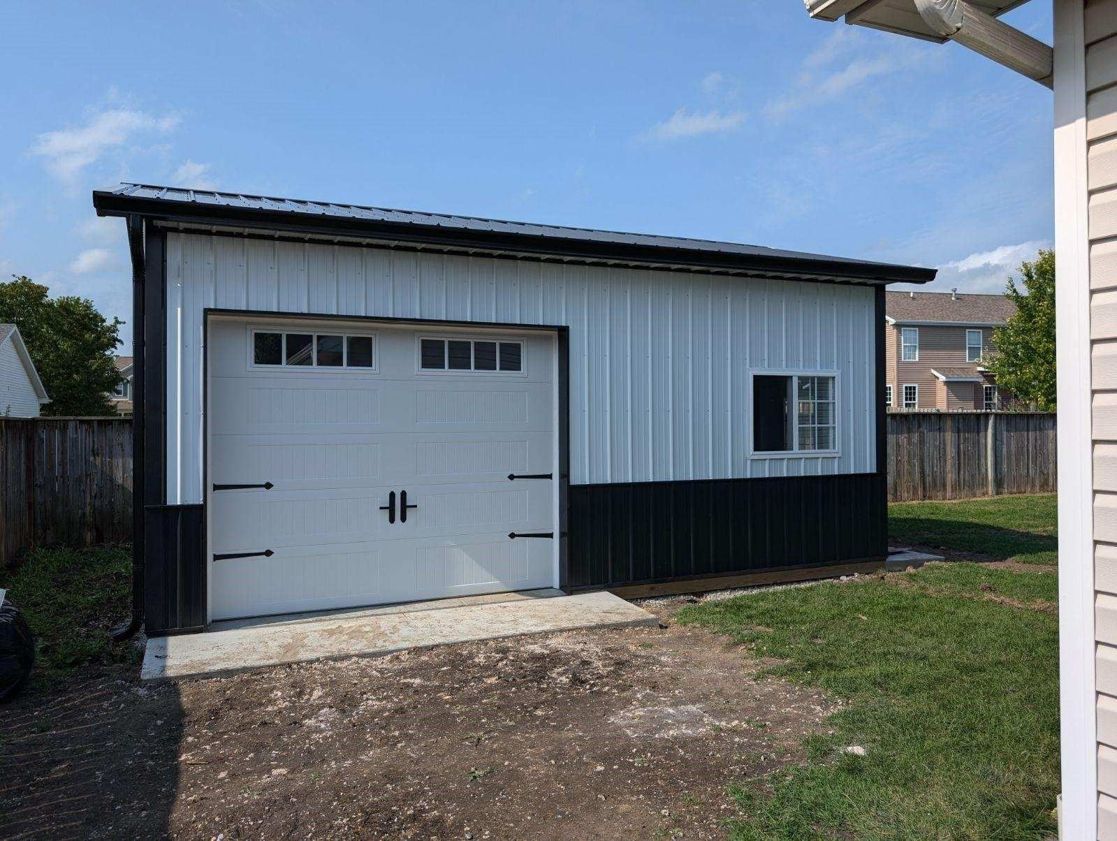 A white and black garage with a black roof is in the backyard of a house.