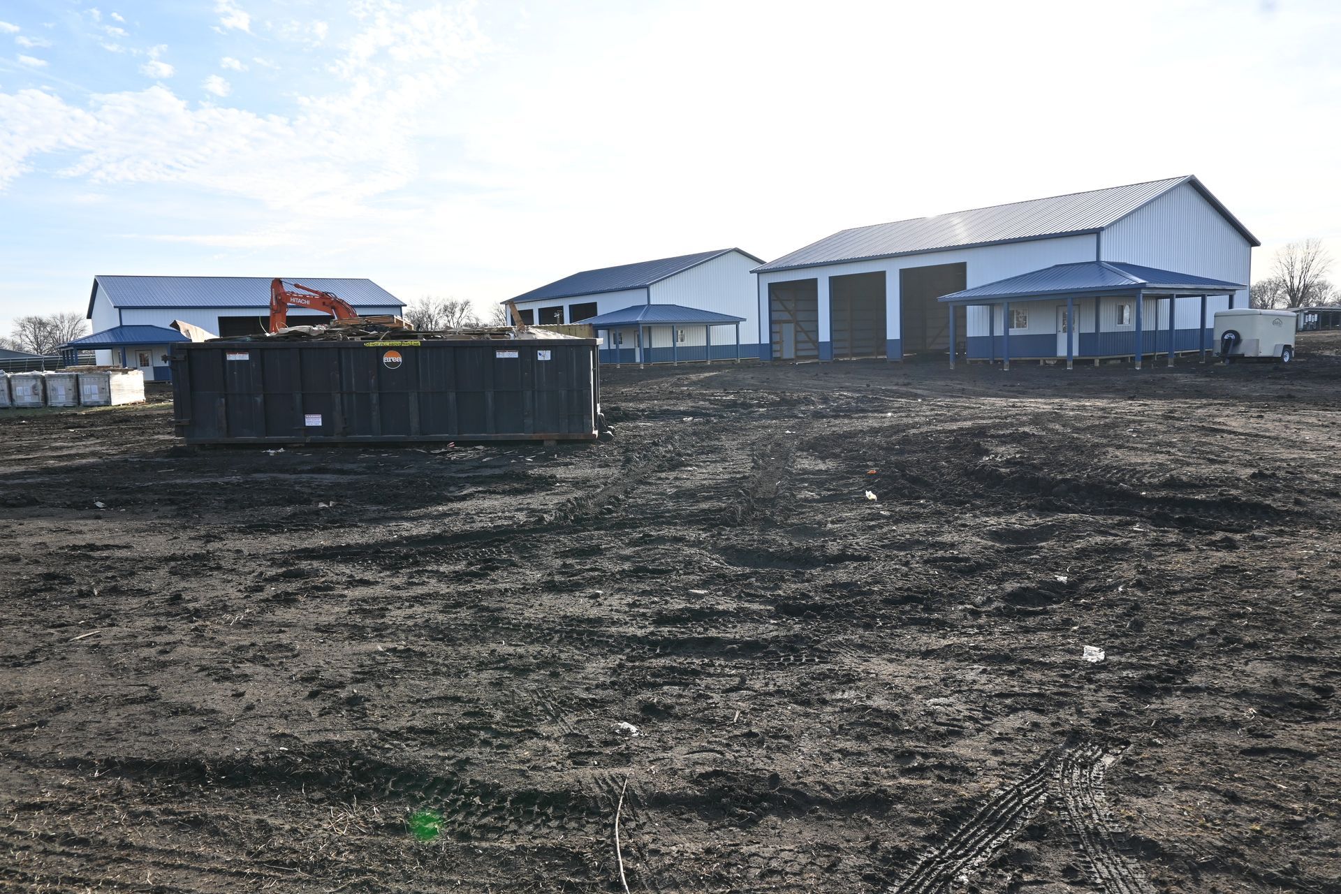 A large black dumpster sits in a dirt field in front of a large building