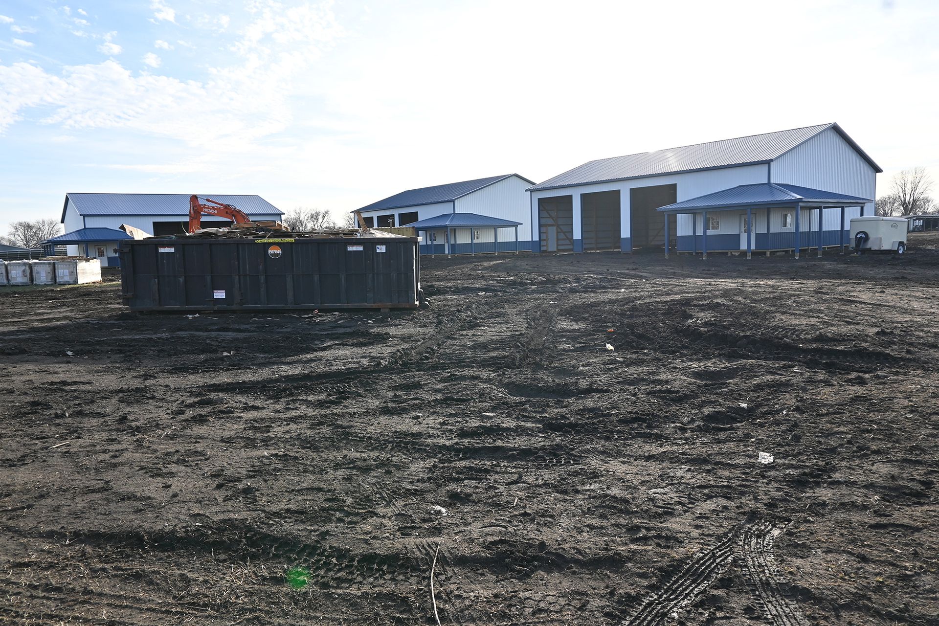 A large black dumpster sits in a dirt field in front of a large building