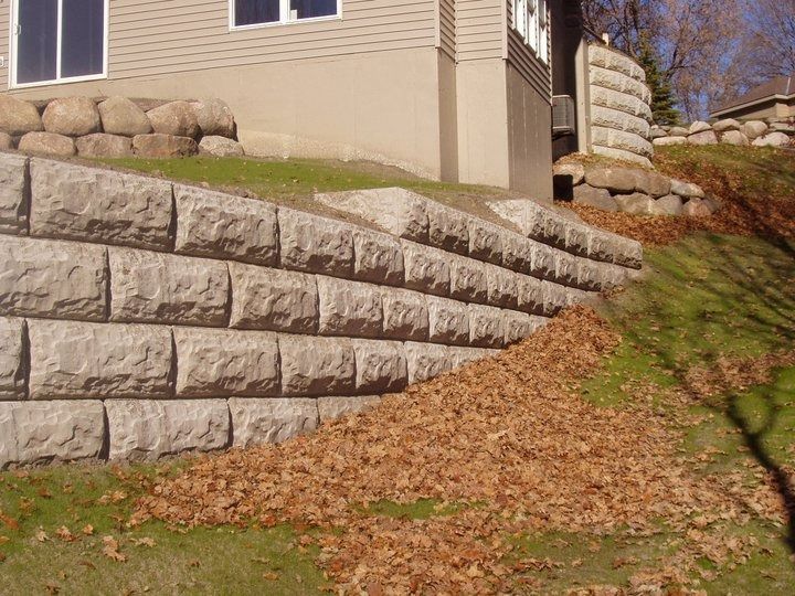 Terraced stone retaining wall beside a beige house with brown mulch and a grassy slope