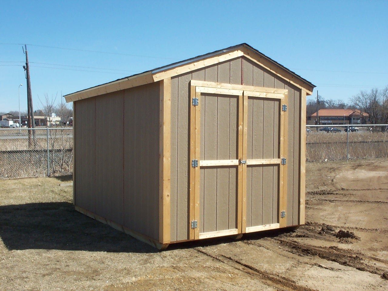 Small tan storage shed with double doors on a dirt lot under a clear sky