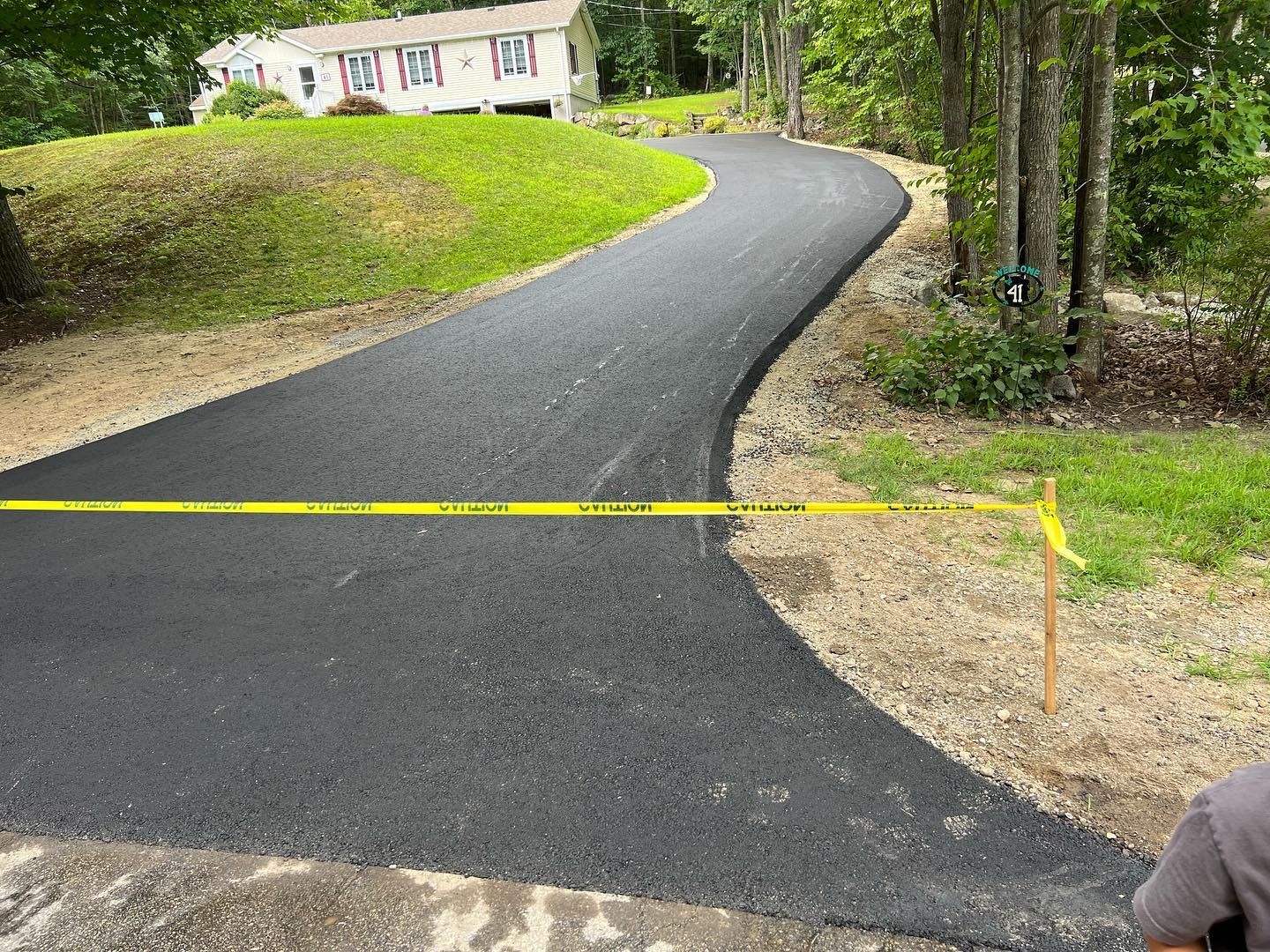 Newly paved asphalt driveway curves uphill towards a house, with yellow caution tape across it.