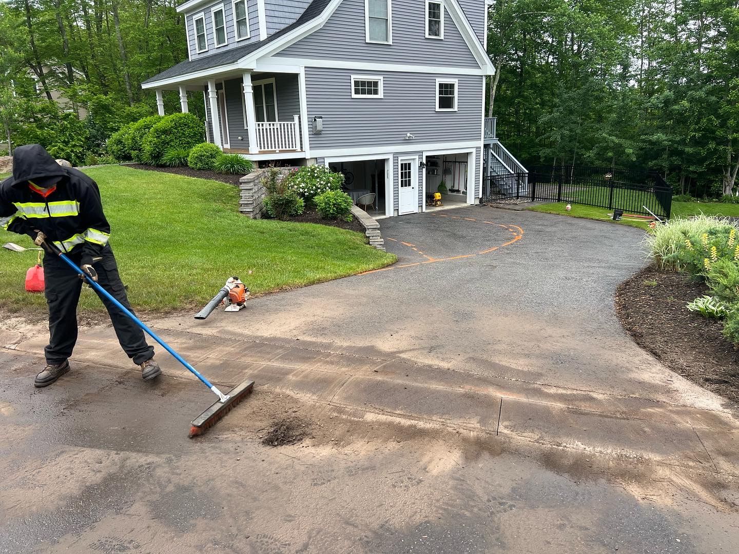 Person sweeping a driveway. A house with a two-car garage is in the background. Green grass and foliage are present.