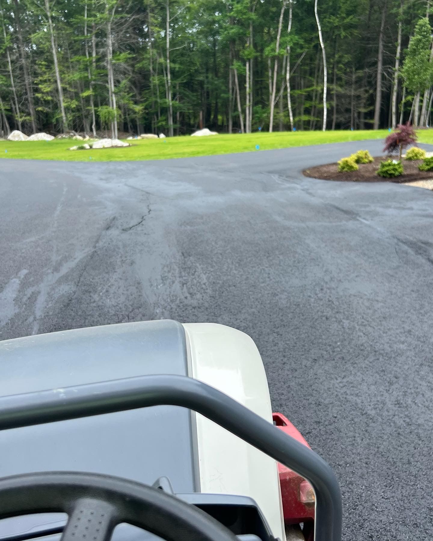 A person on a riding lawnmower on a dark paved driveway, trees in the background.