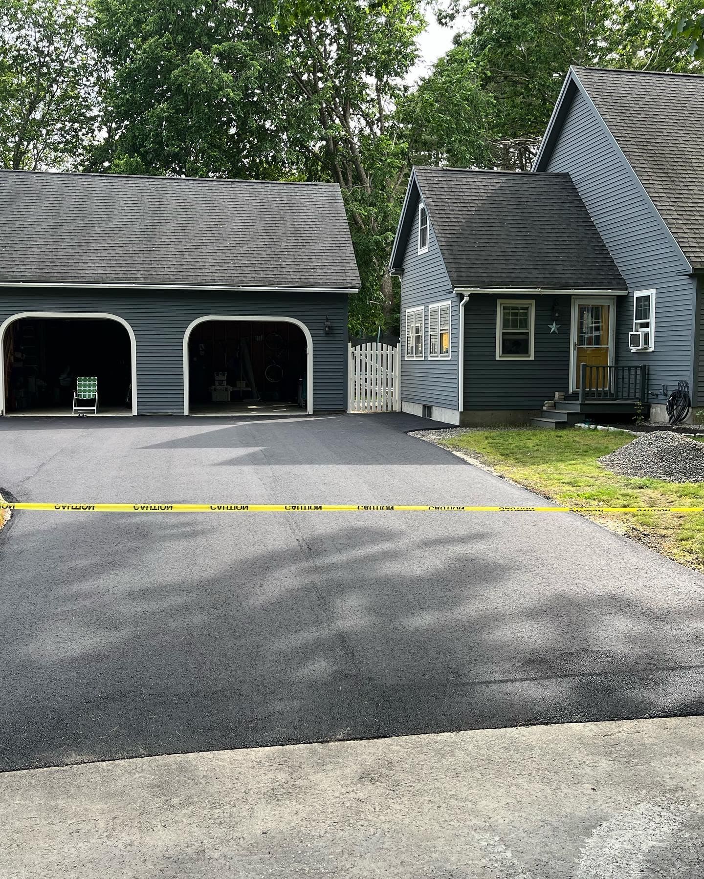 Asphalt driveway leading to a blue house and garage, connected by a white gate. Yellow caution tape is across the driveway.