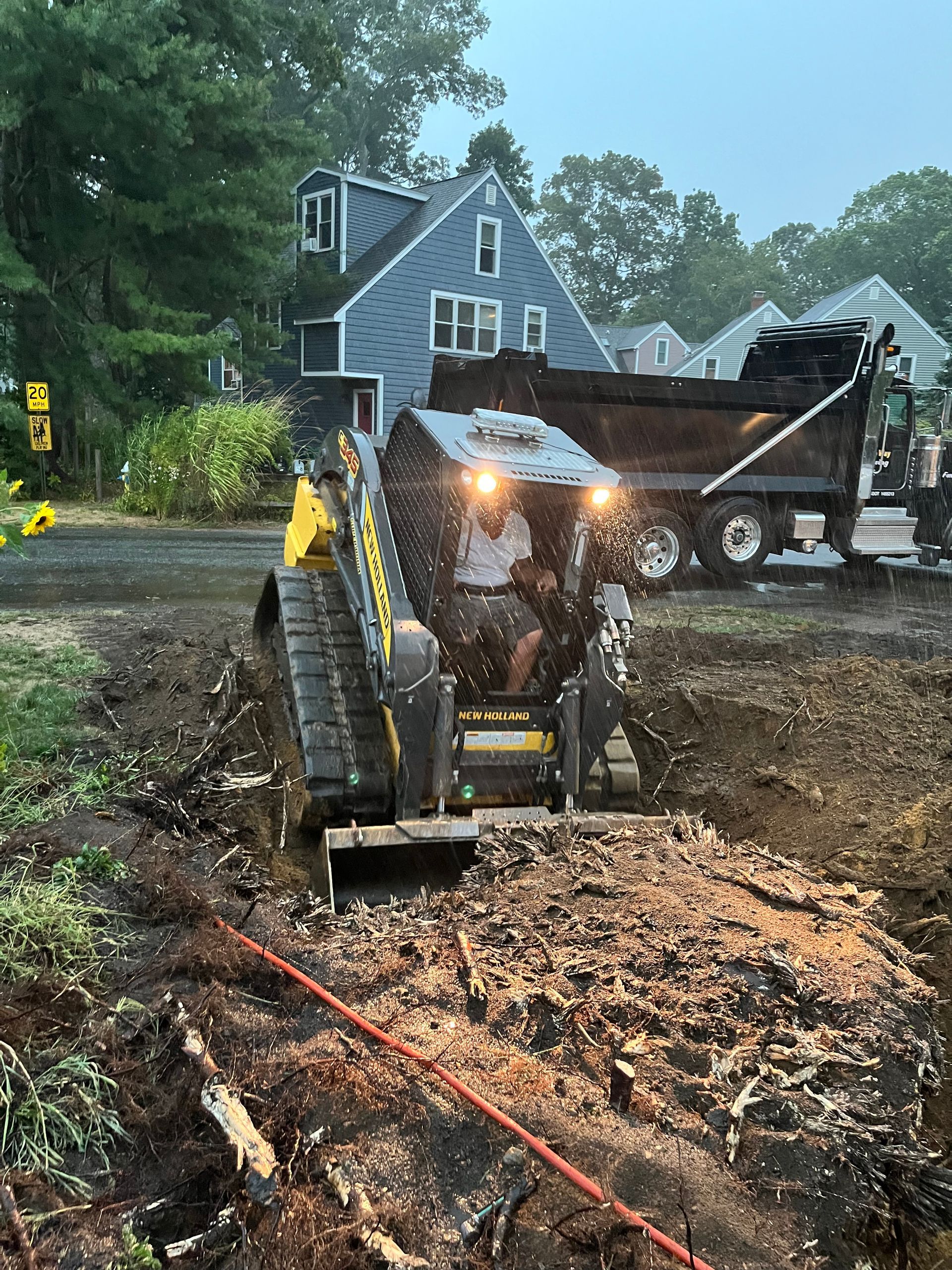 A person operates a skid steer to dig in a muddy area near a residential street, with a dump truck in the background.