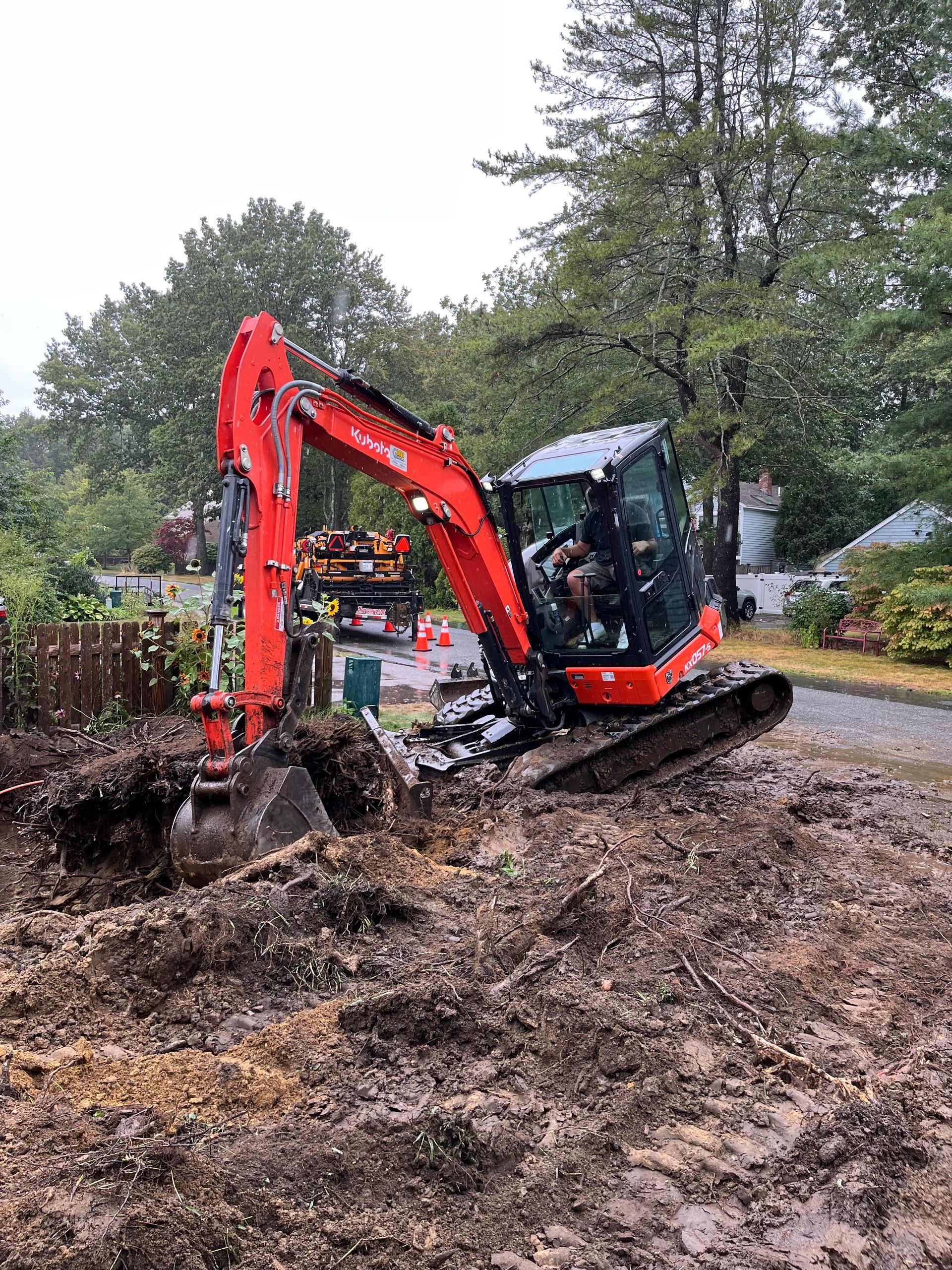Red excavator digging in muddy soil near a road and fence, with a person inside.