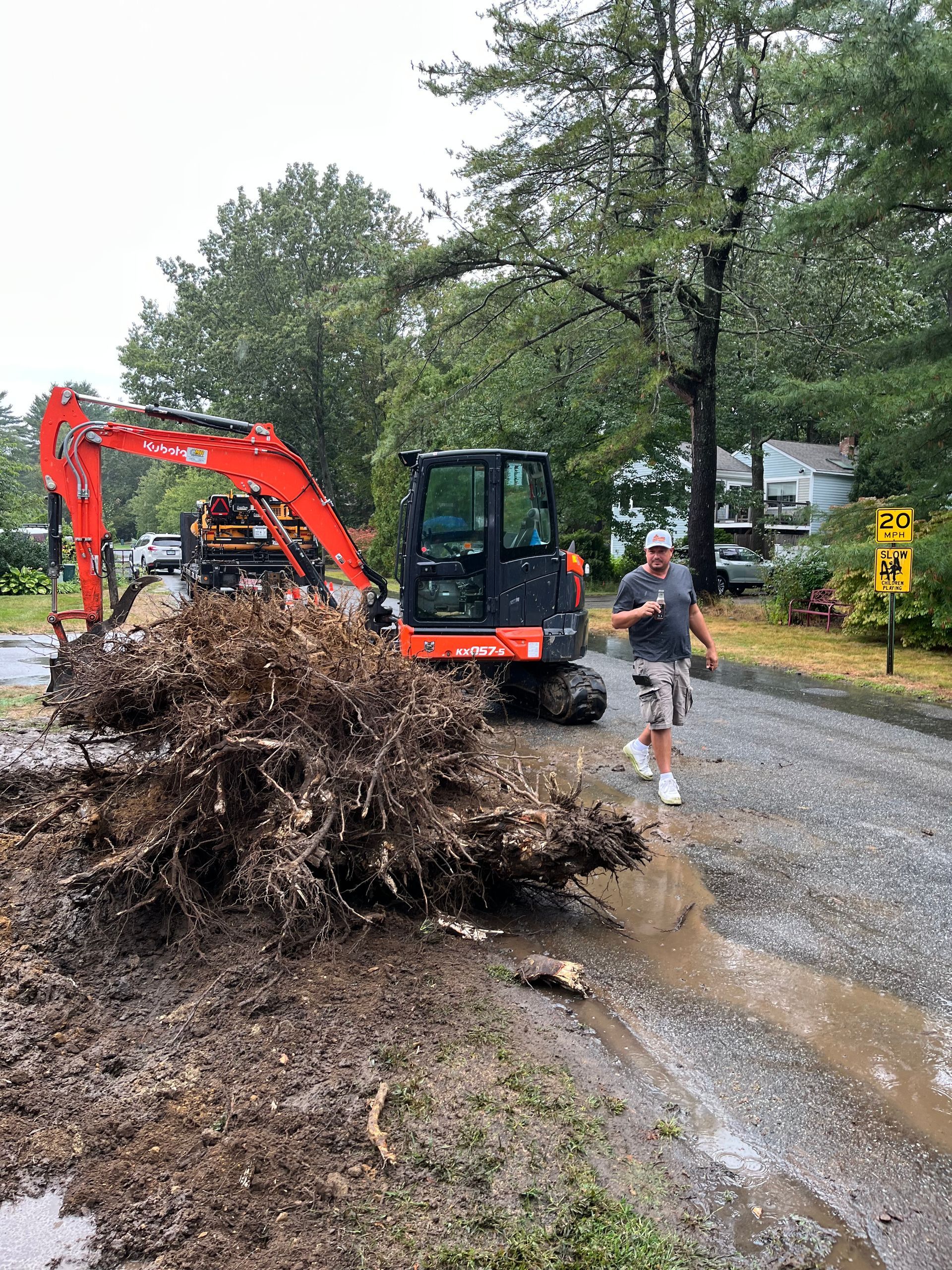 Excavator next to a large uprooted tree on a wet road; a person runs by the scene.