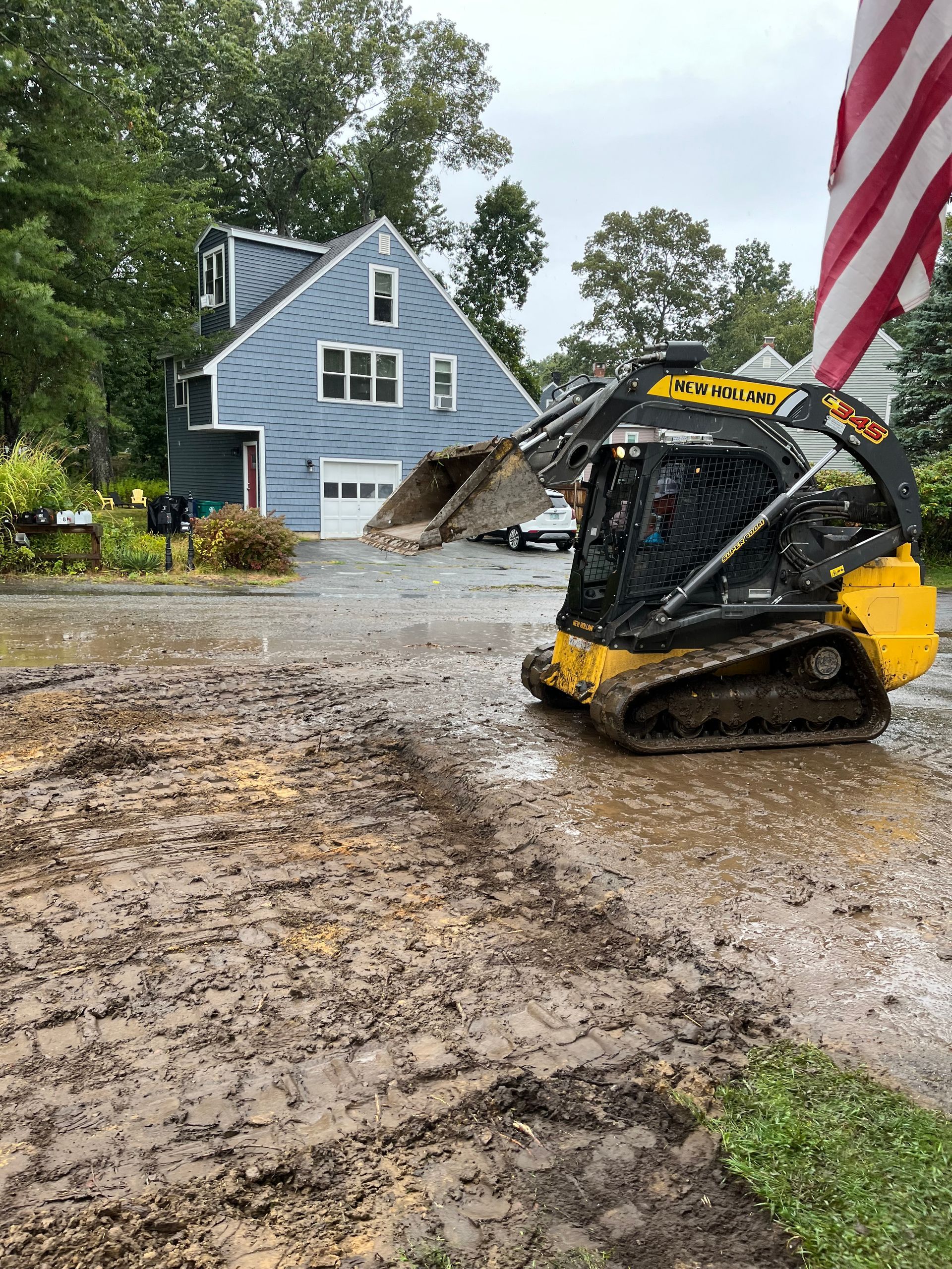 A small excavator on a muddy driveway in front of a blue house, with a waving American flag.