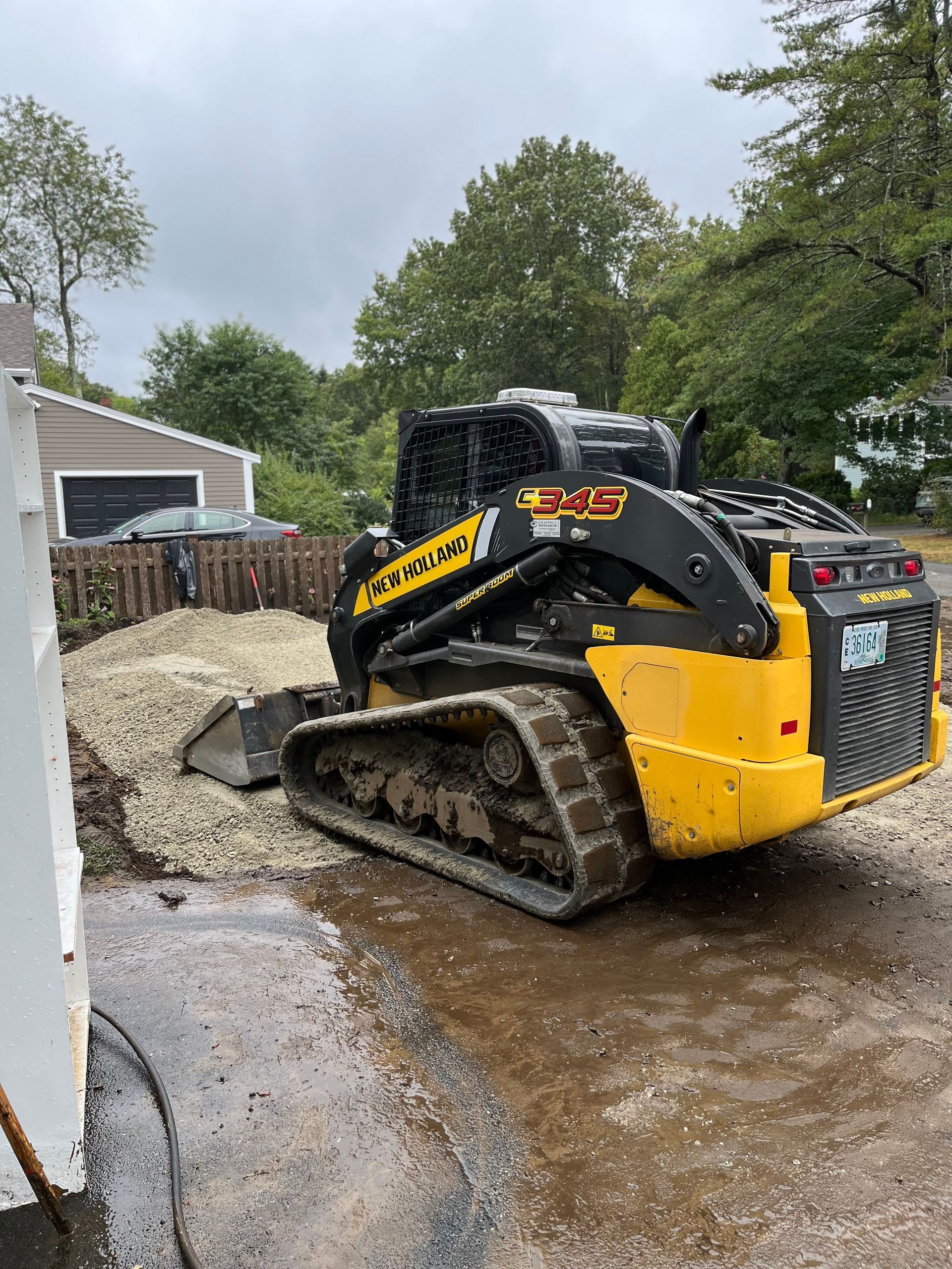 Yellow skid steer loader next to gravel pile, parked on a wet driveway.