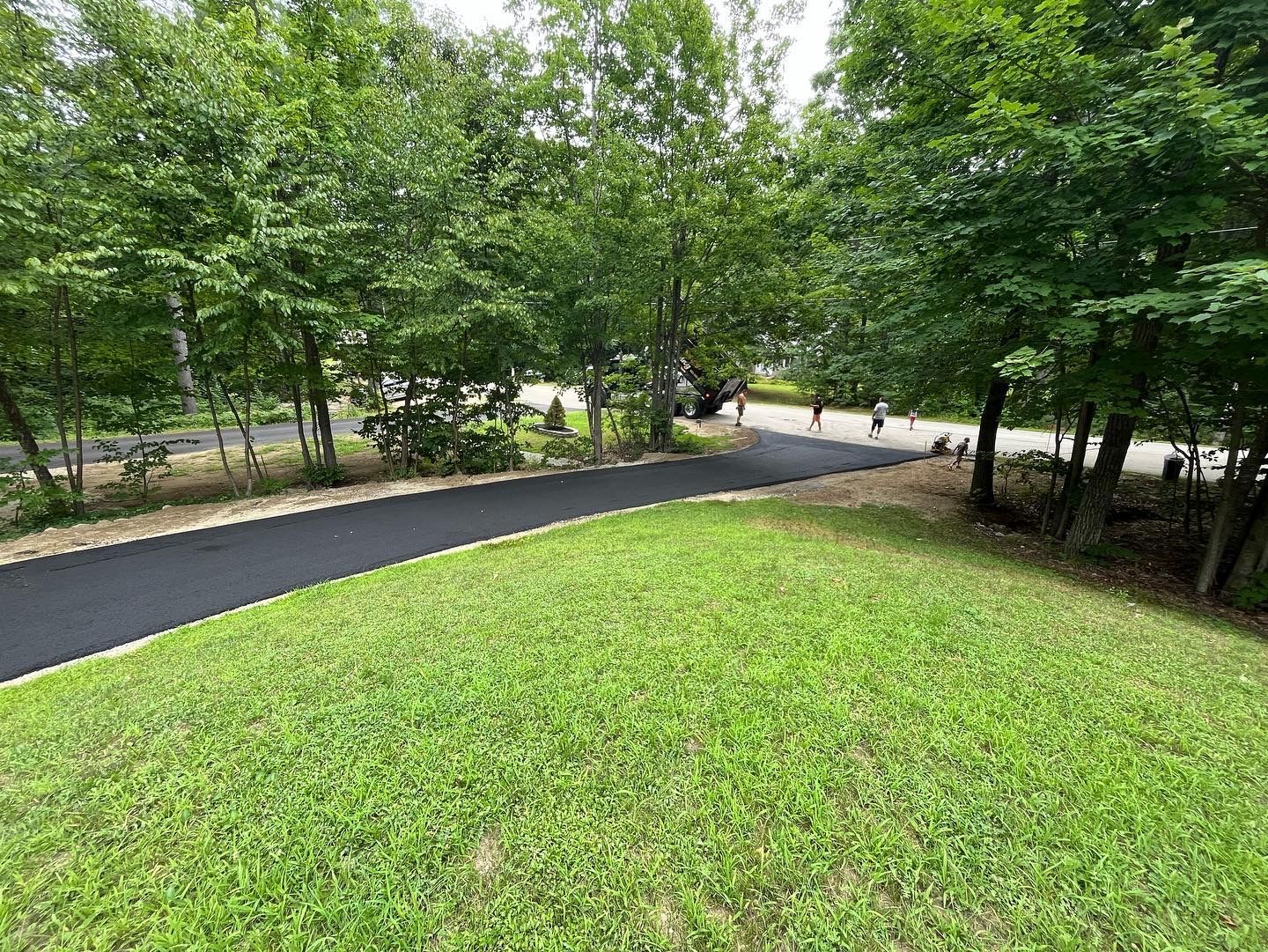 A paved walking path curves through a grassy area lined with trees; people are seen further down the path.