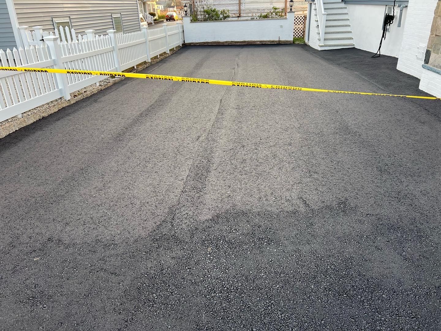 Newly paved driveway with yellow caution tape, white fence, and stairs.