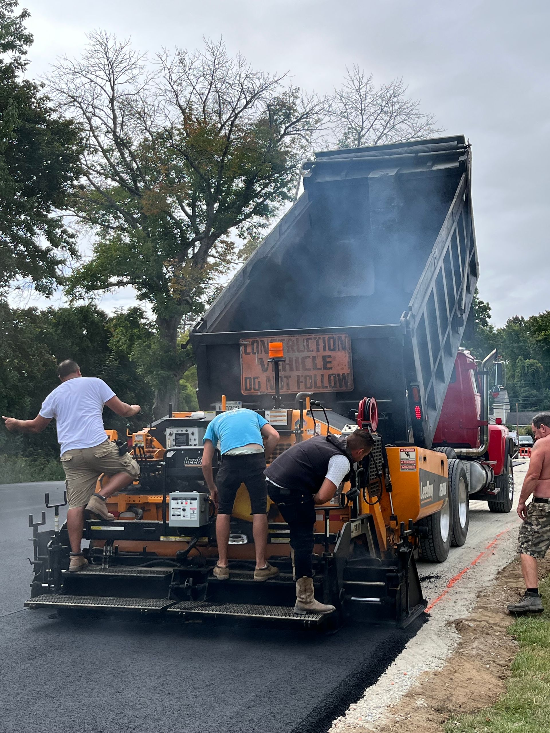 Asphalt paving crew at work: dump truck unloading into the paver, workers leveling, on a roadway.