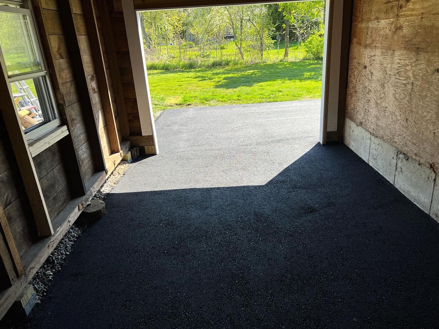 Asphalt driveway inside an open garage with exposed wooden framing and a view of grass and trees.