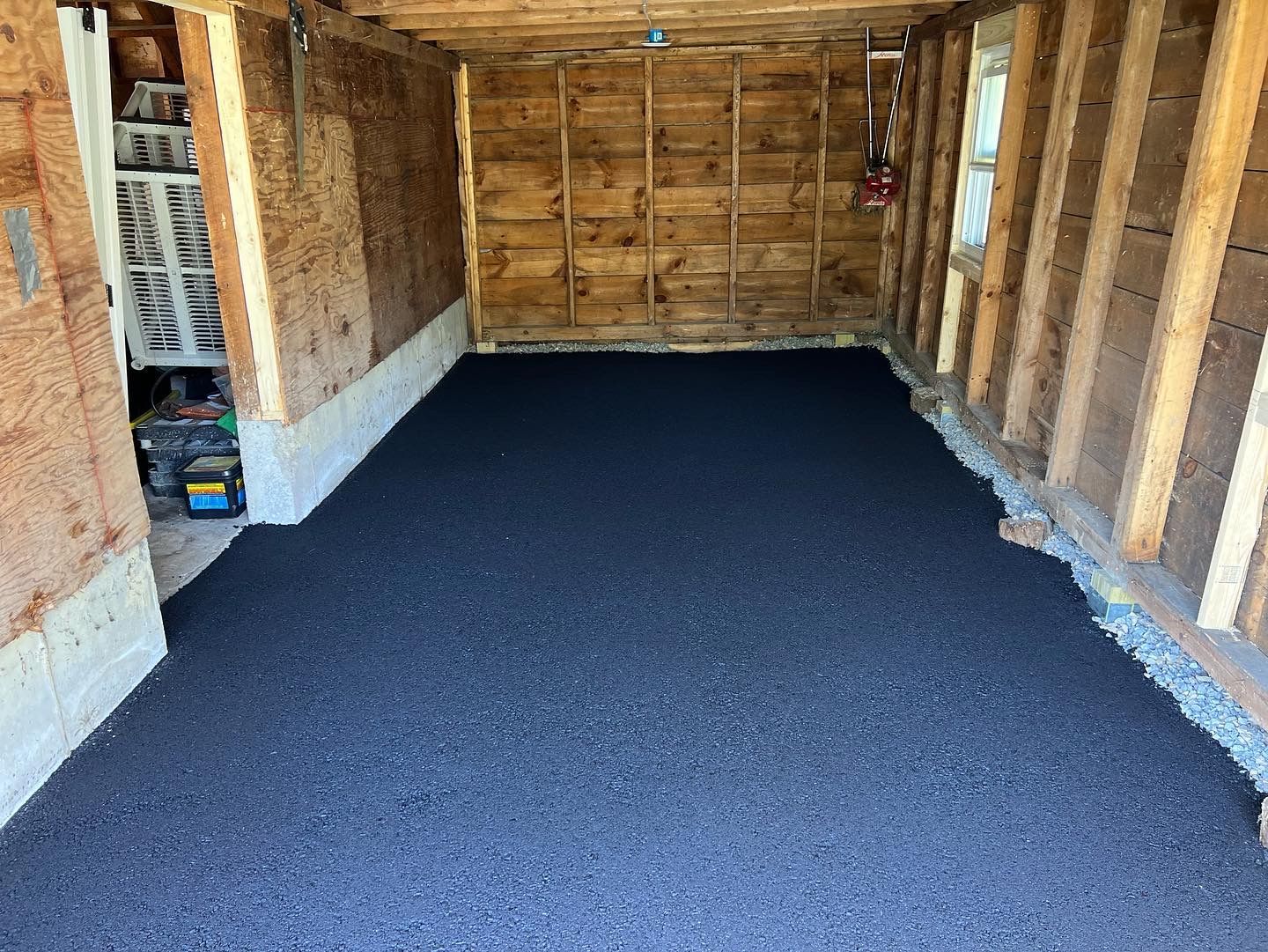 Inside of a garage; newly paved dark gray asphalt floor. Wooden walls and support beams.