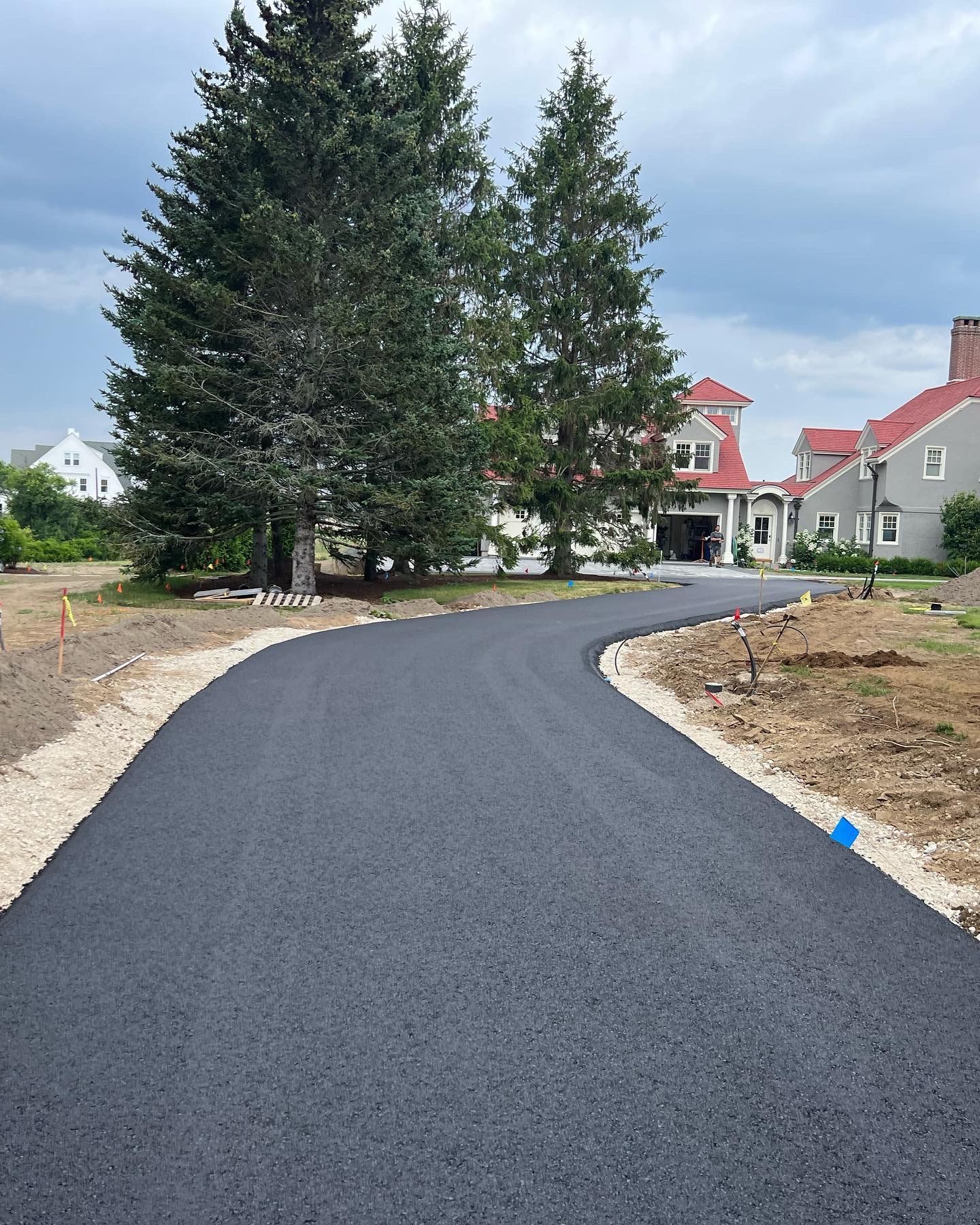 Newly paved asphalt driveway curves towards a house with tall trees in the background. Cloudy sky.