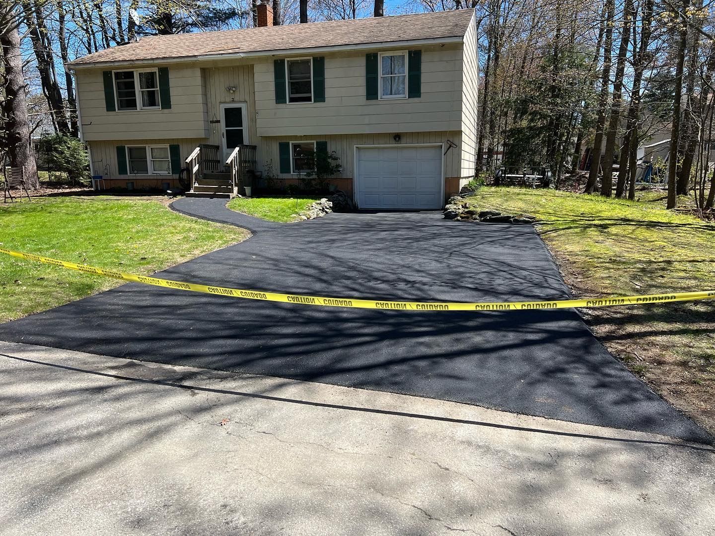 Newly paved driveway in front of a two-story beige house with green shutters, blocked by yellow caution tape.