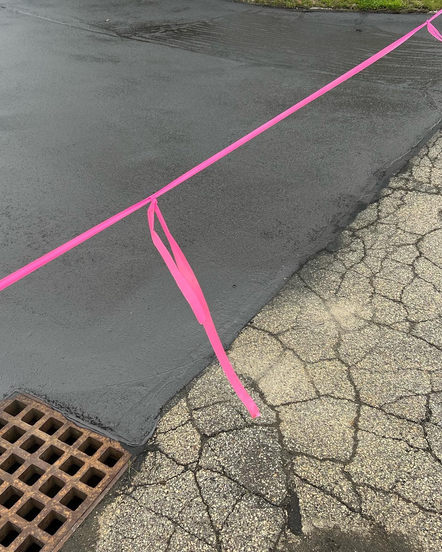 Pink utility marking tape across asphalt and cracked pavement next to a drain.