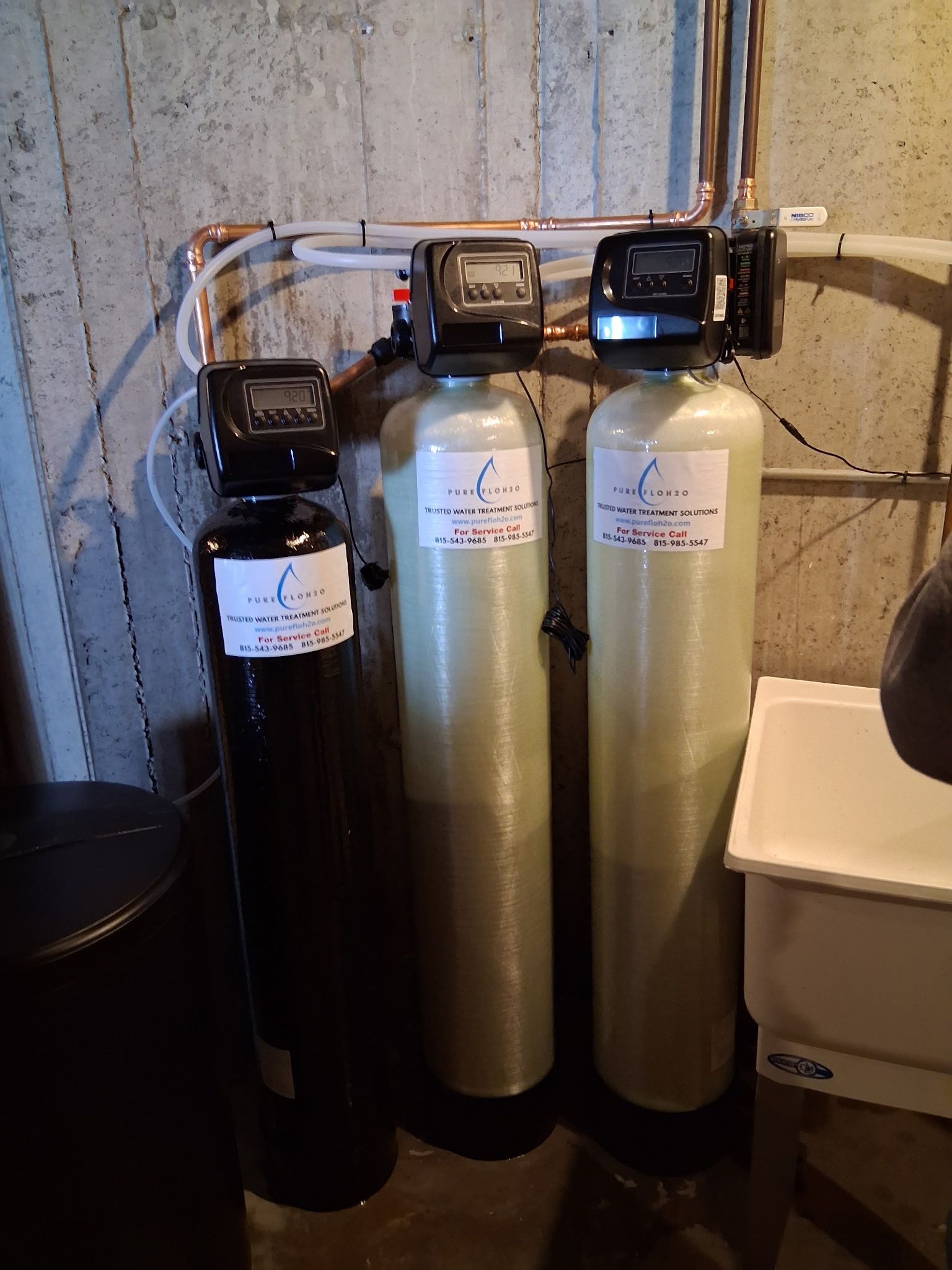 Three water filtration tanks with digital control heads are installed against a concrete wall near a utility sink. 