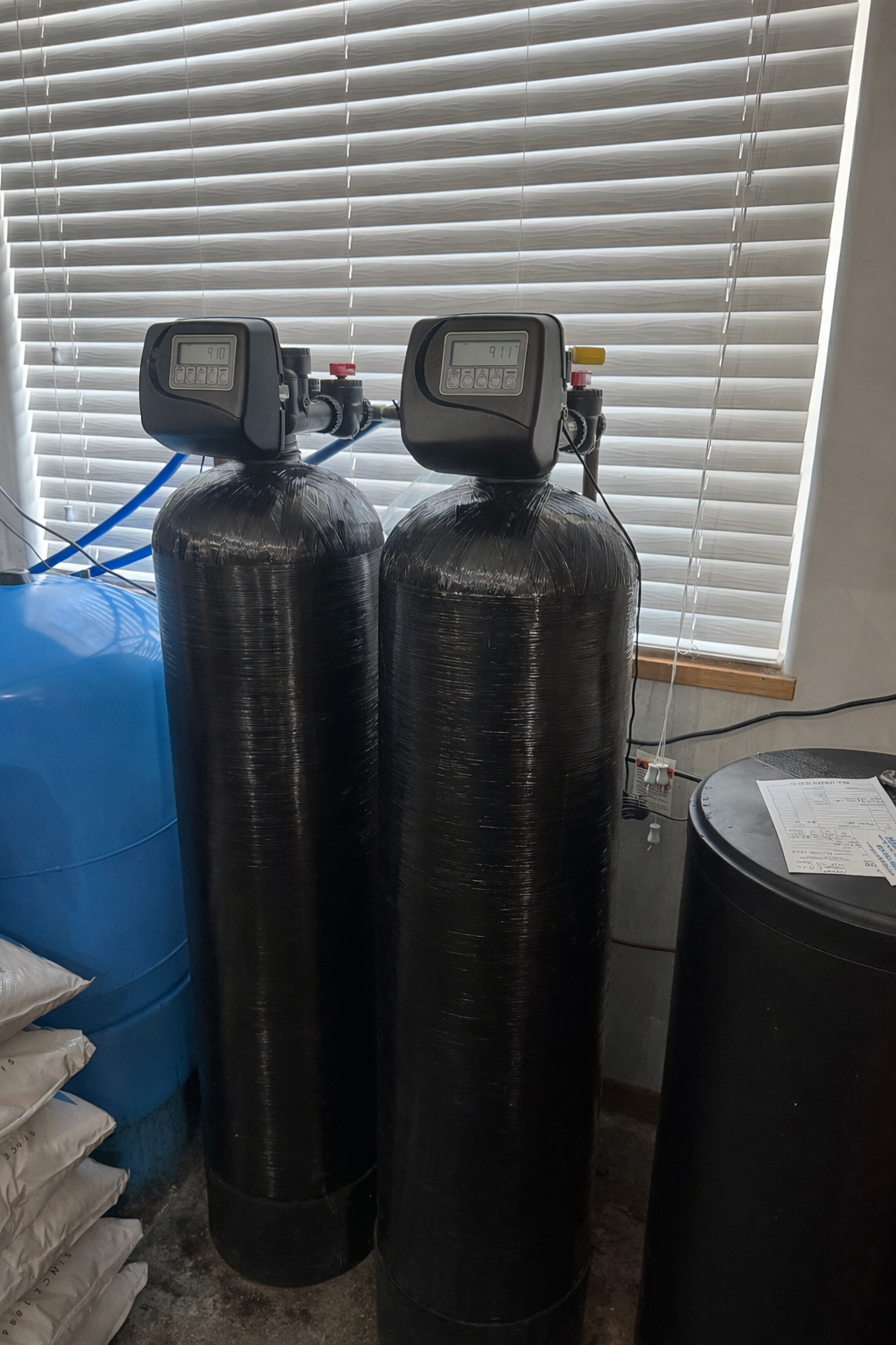 Two tall black water filtration tanks with digital displays, next to a blue tank and a black barrel.